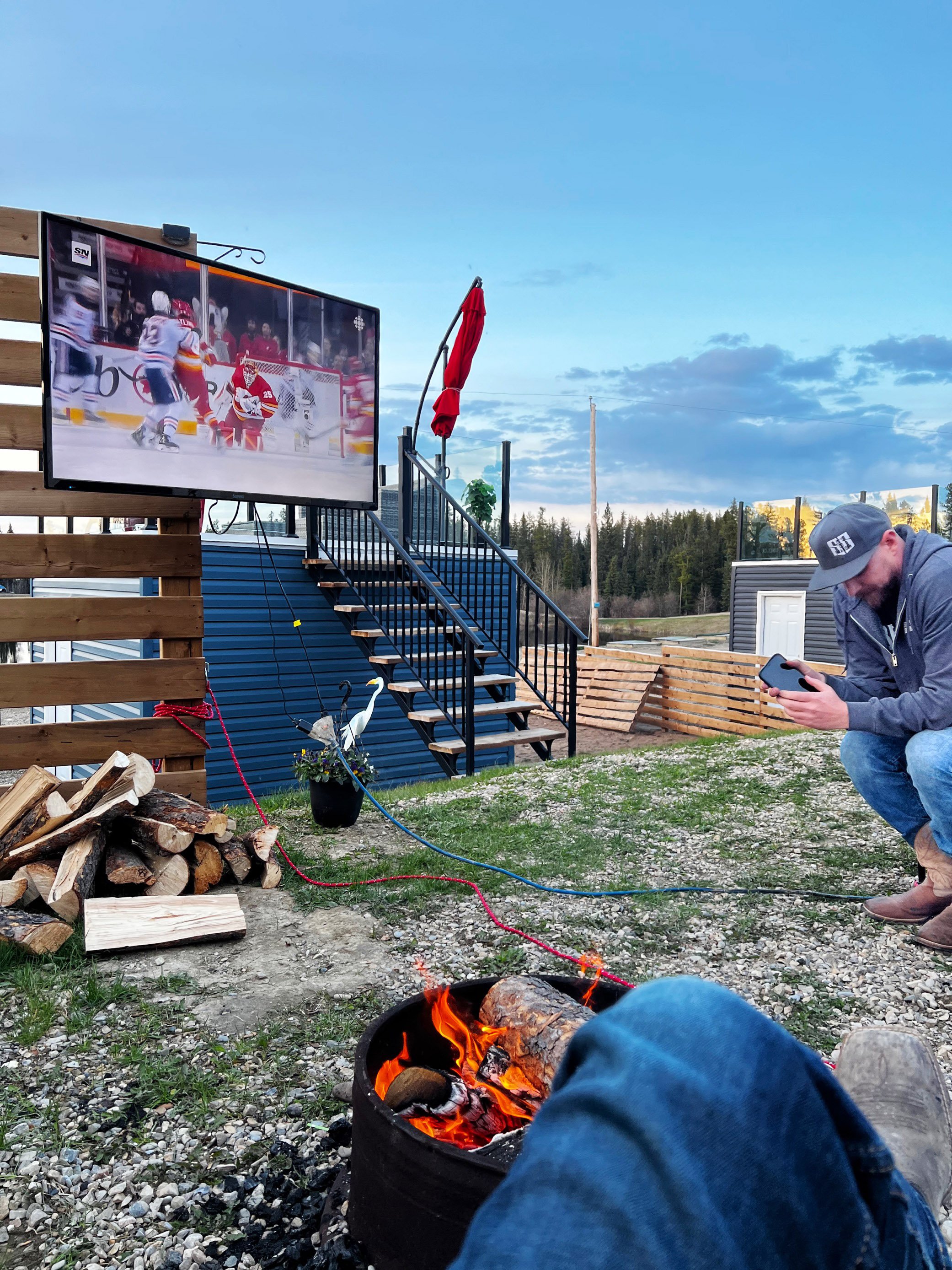 Outdoor scene with a fire pit, stacked firewood, and a man squatting while looking at his phone. A large flat-screen TV displays an hockey game, mounted on a wooden structure. A red umbrella is closed, and there are trees and a blue sky with clouds i
