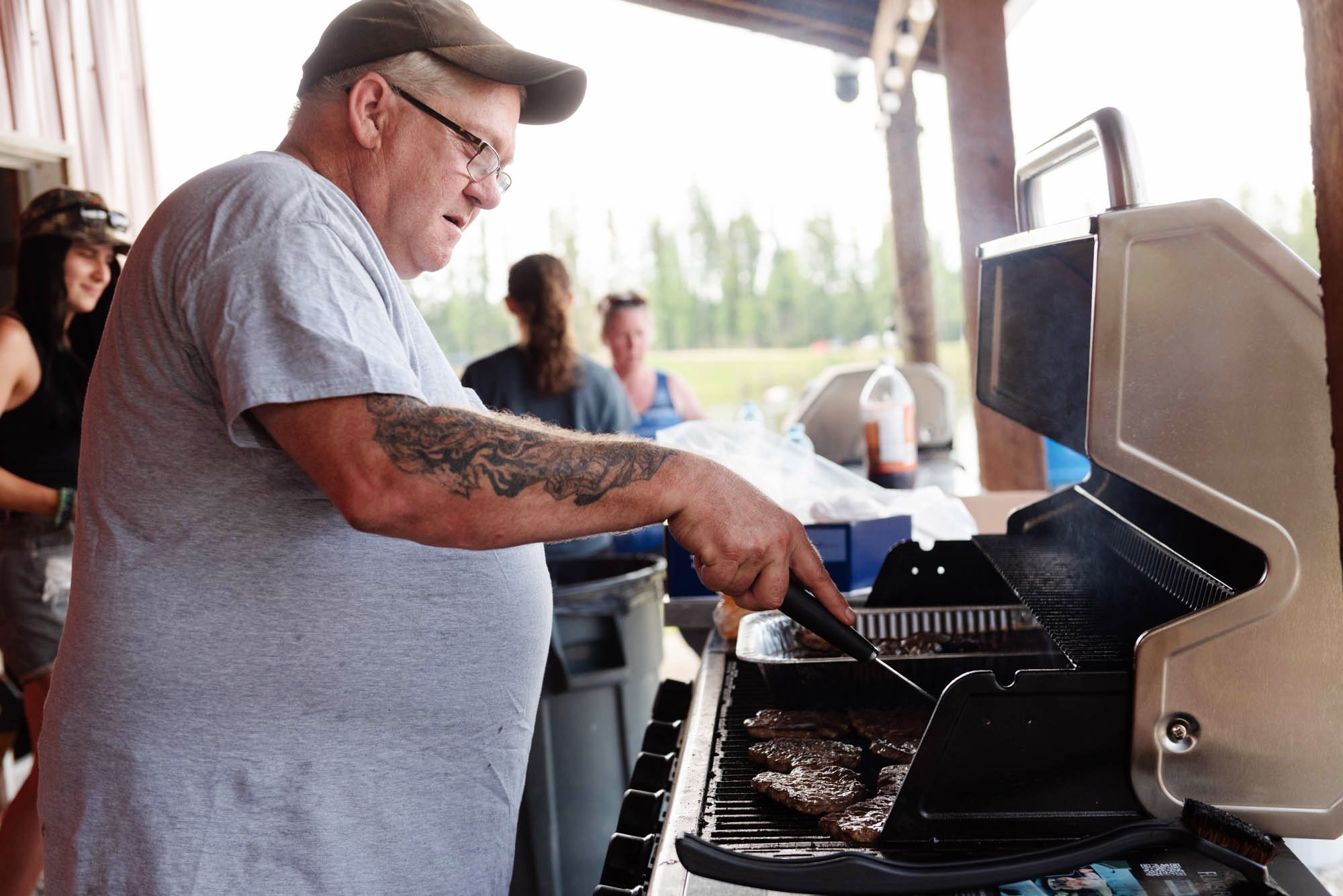 Man with glasses and tattoos cooking burgers on an outdoor grill, with a group of people in the background.
