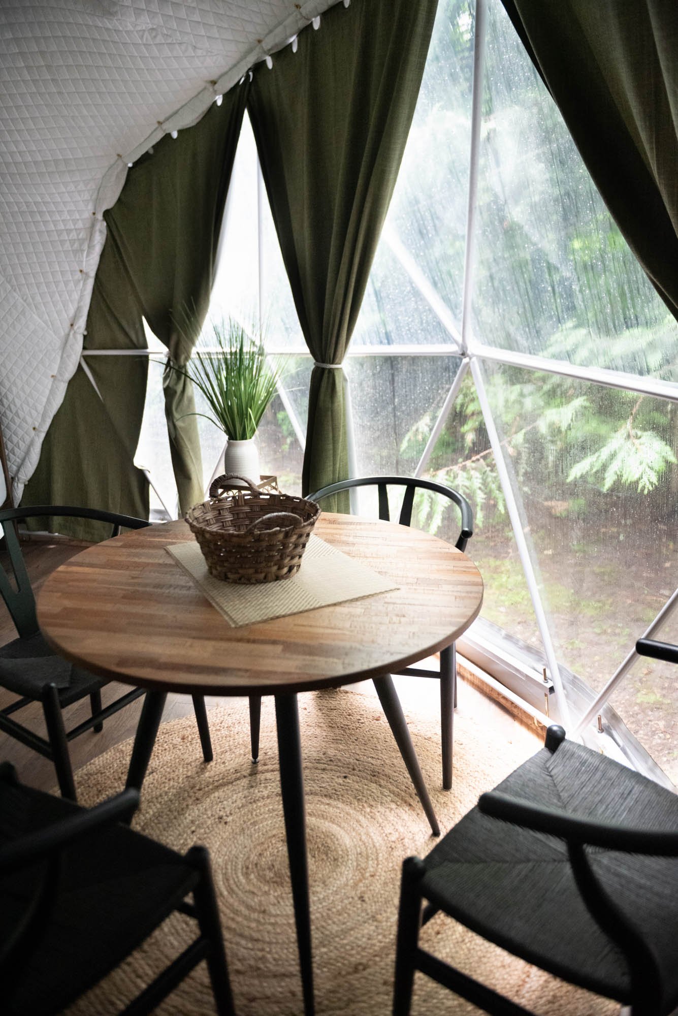 Inside a cozy bubble tent with black curtains, a wooden table with a woven basket, black chairs, a potted plant, and a window showing a garden outside during rain.