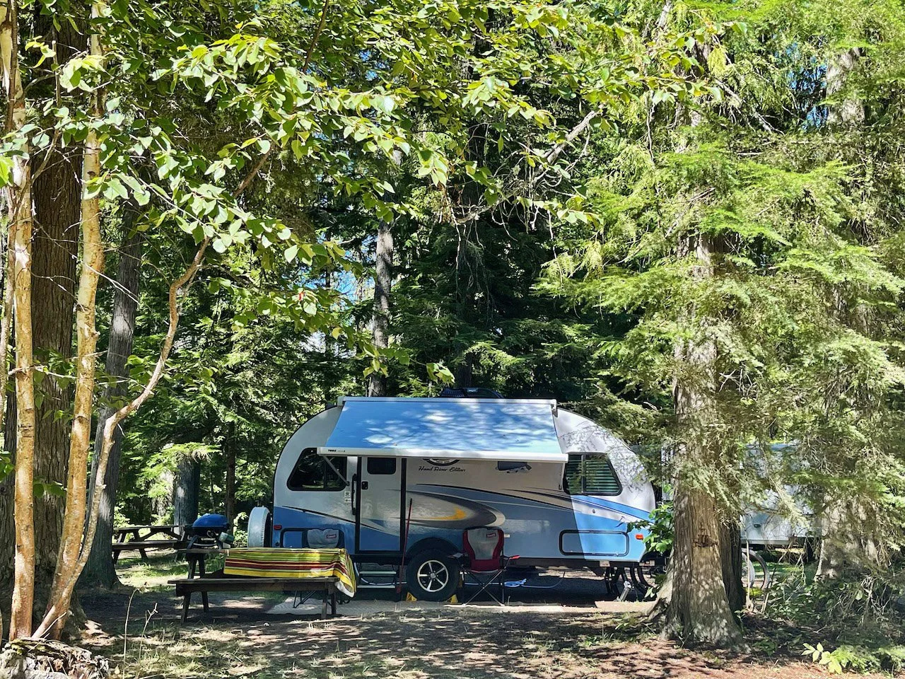 A blue and gray camper trailer parked in a forested area with tall trees and sunlight filtering through the branches. There is a picnic table with a yellow-striped tablecloth and a folding chair nearby.