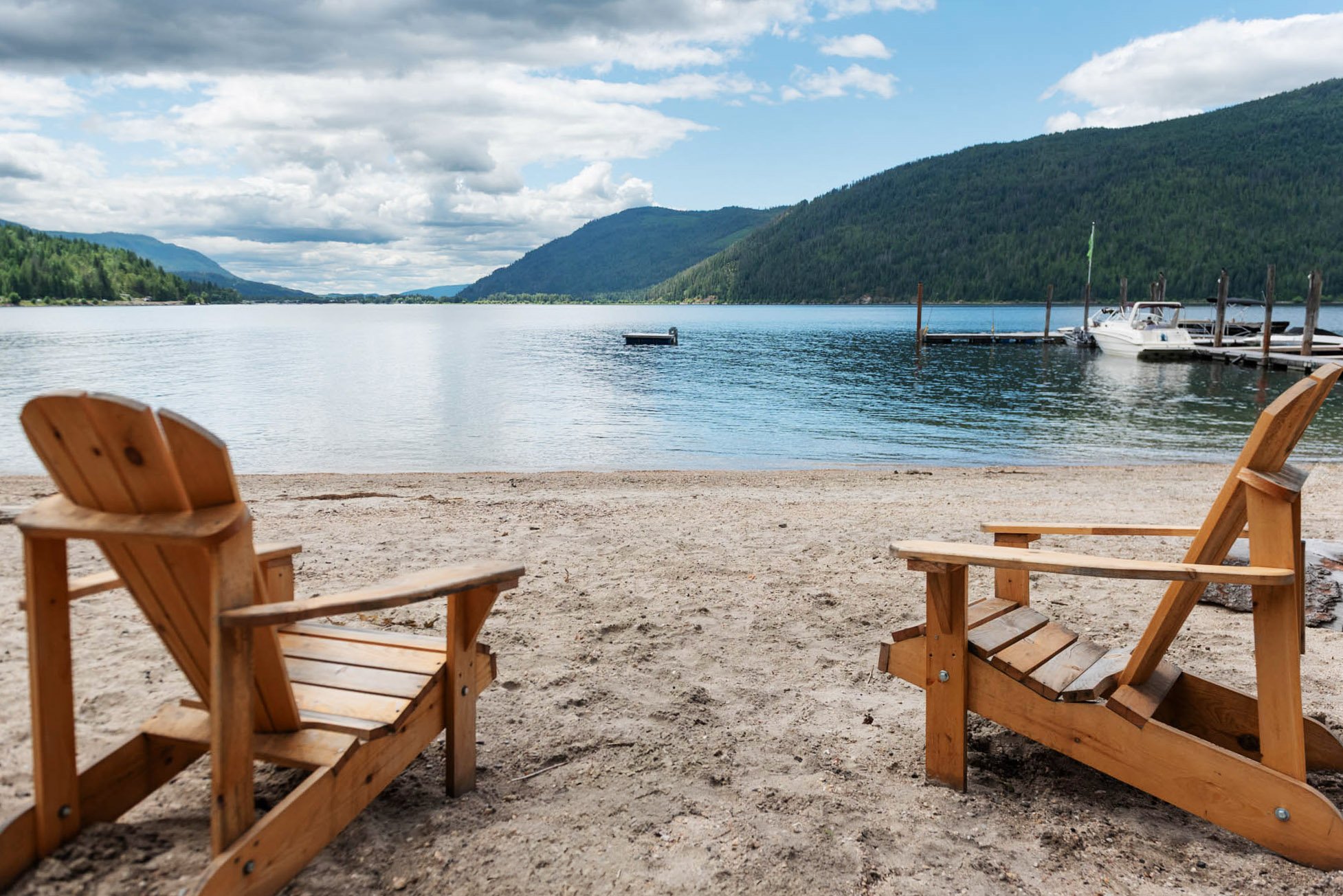View of a lake with hills in the background, seen from a sandy beach area with two wooden chairs in the foreground, and a dock with boats on the right side.