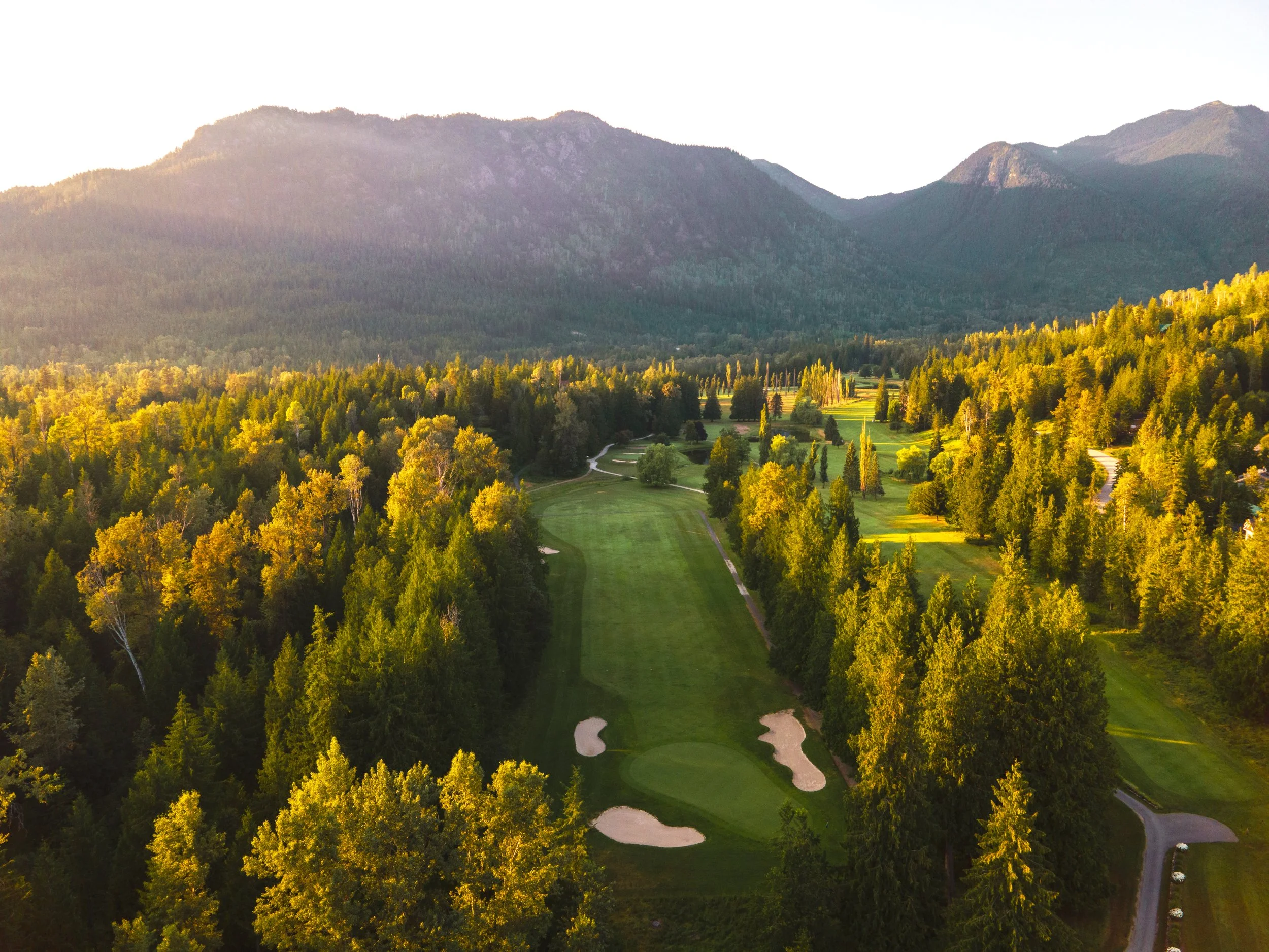 A scenic view of a golf course surrounded by trees with mountains in the background, bathed in golden sunlight.