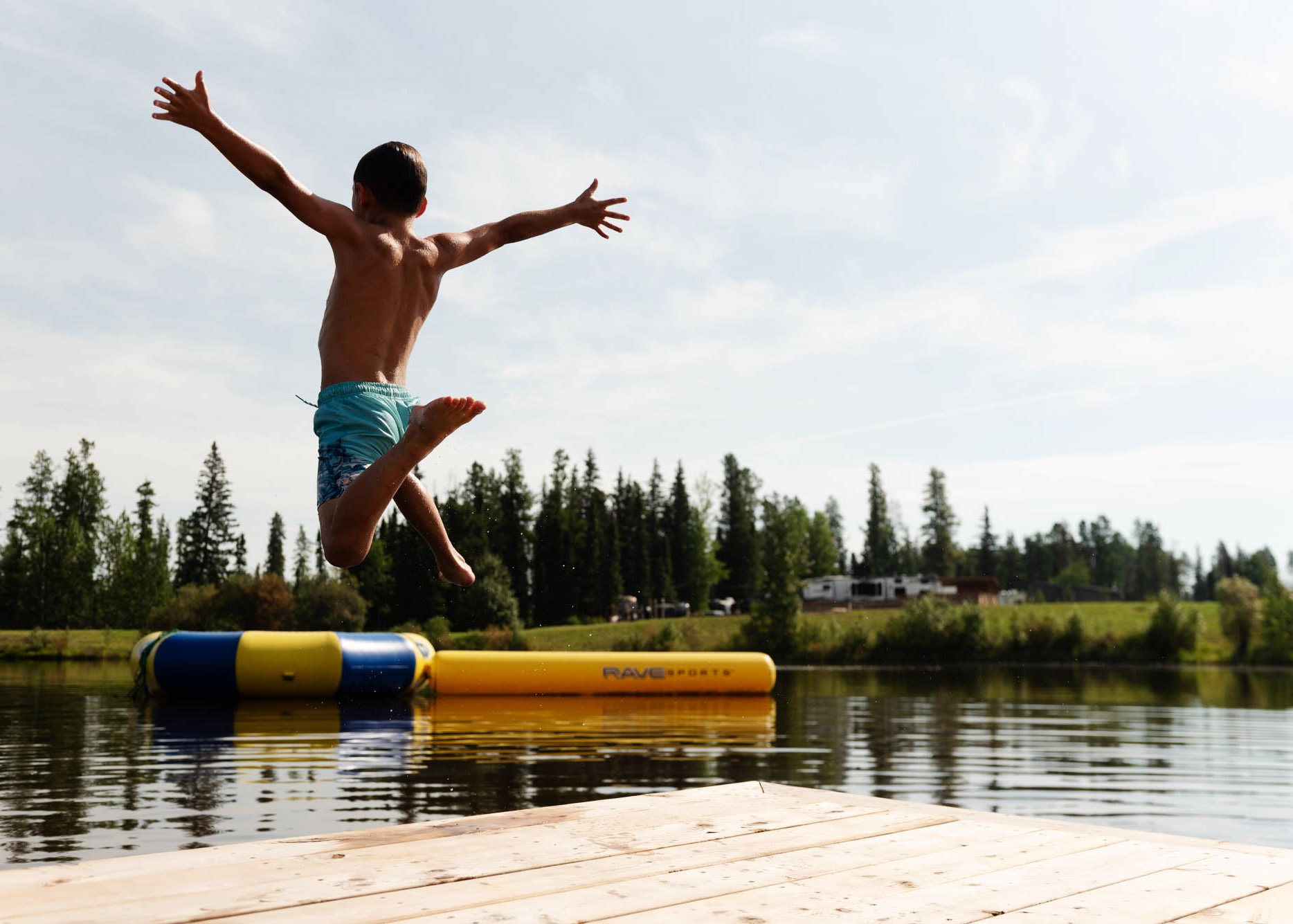 Boy jumping off a floating dock into a lake with a water toy, surrounded by trees and a partly cloudy sky.