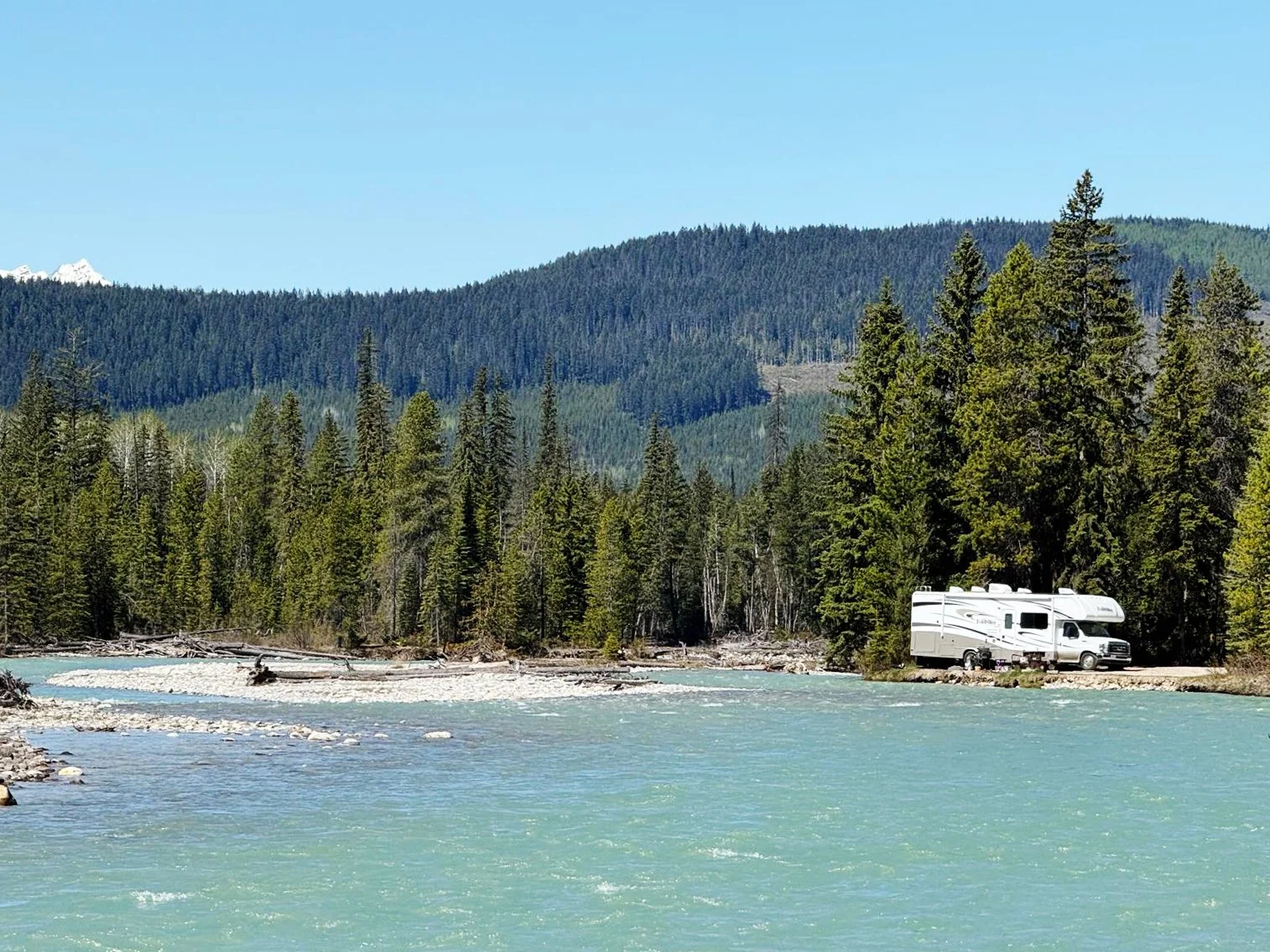A white RV parked on a small patch of land beside a river, surrounded by dense green forest and mountains in the background under a clear blue sky.