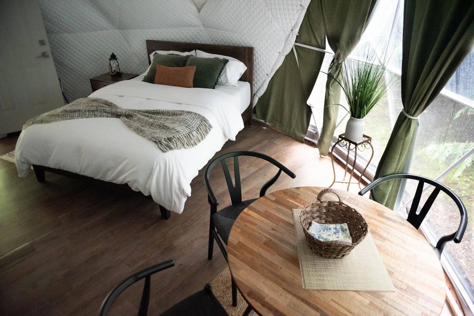 A cozy bedroom with a bed, green curtains, a potted plant by the window, and a wooden table with black chairs.