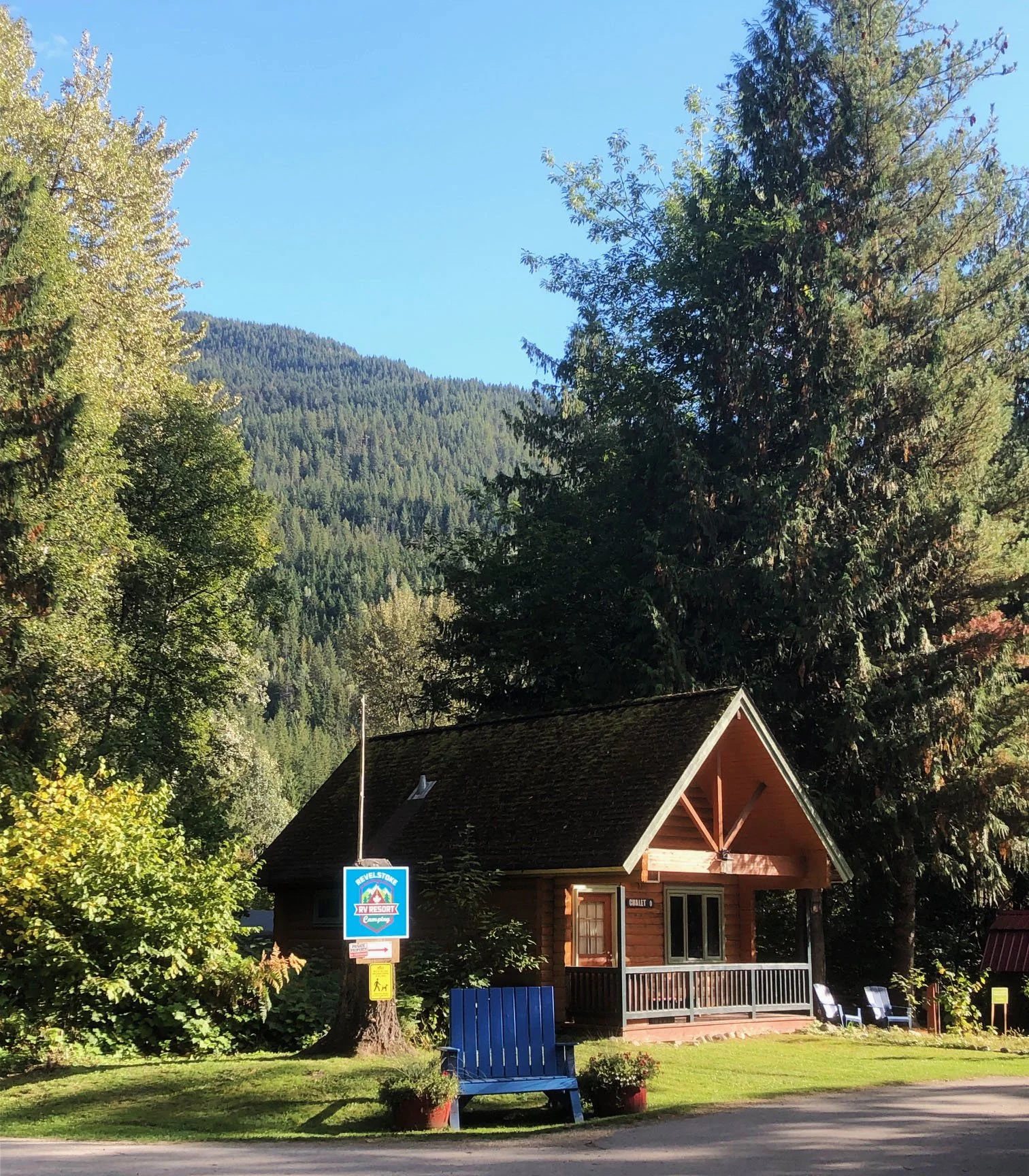 A small wooden cabin with a porch, surrounded by green trees and mountains in the background, under a clear blue sky.