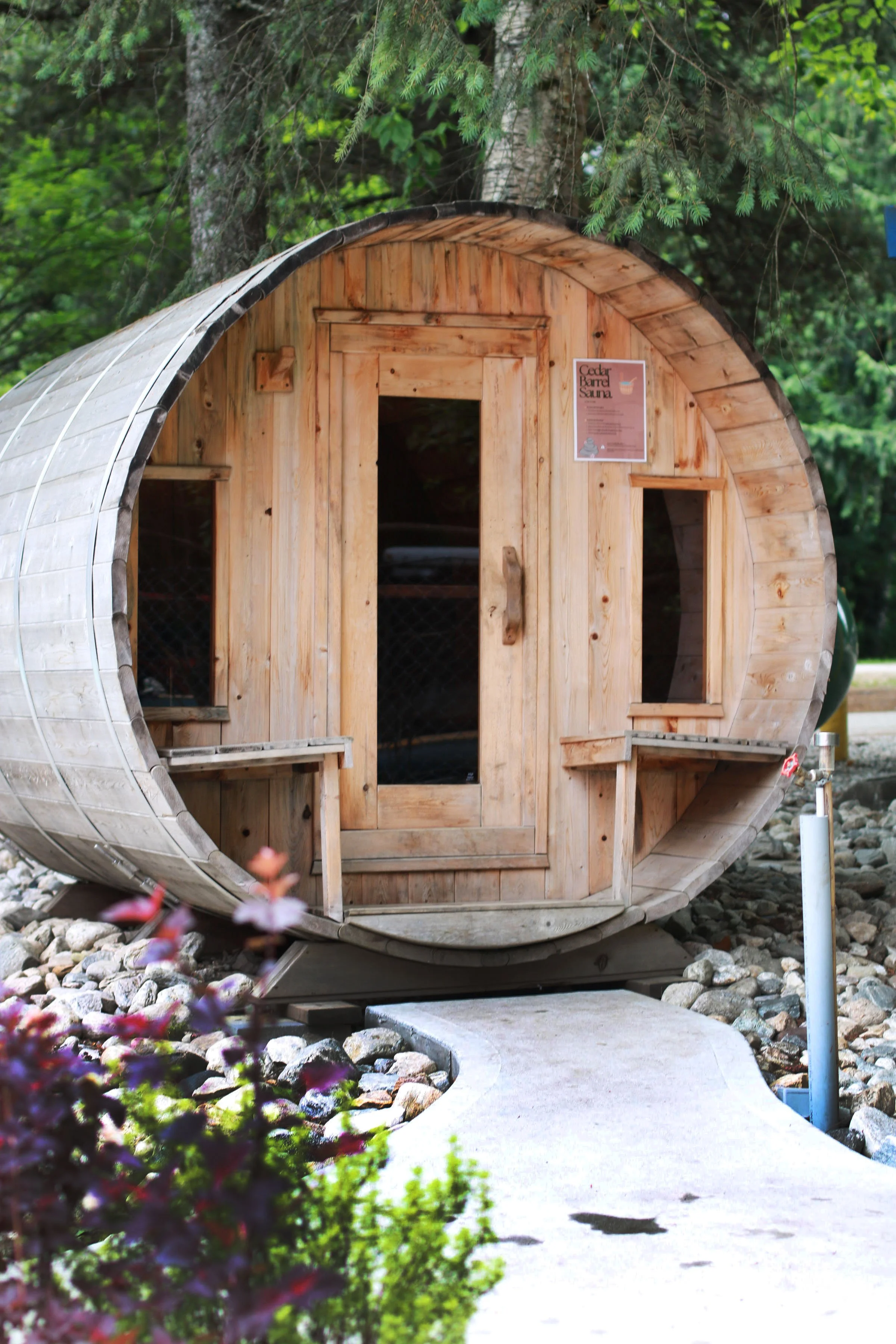 A small wooden cedar barrel sauna with a curved design surrounded by greenery and rocks, with a pathway leading to the entrance.