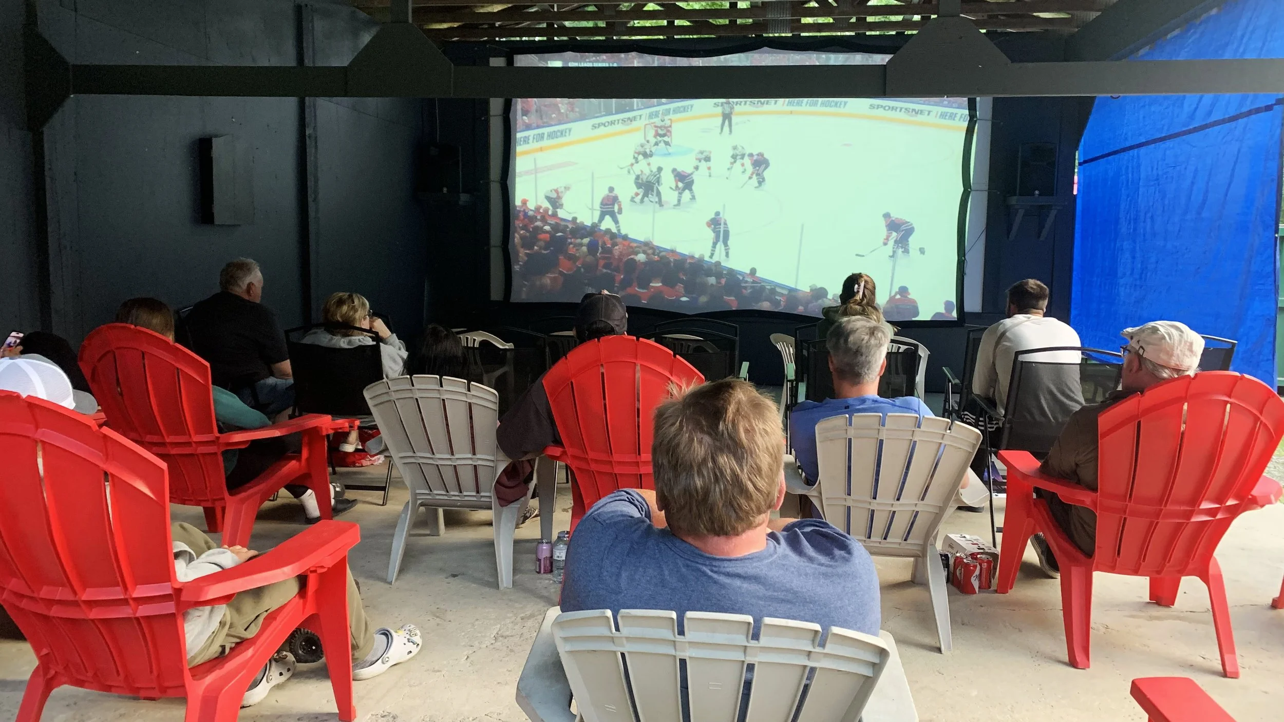 People sitting on red and white chairs watching a hockey game on a large screen in an outdoor setting.
