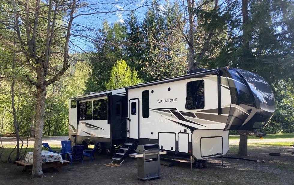 A white and black Avalanche fifth wheel RV parked in a wooded campsite with a picnic table, chairs, and outdoor grill.