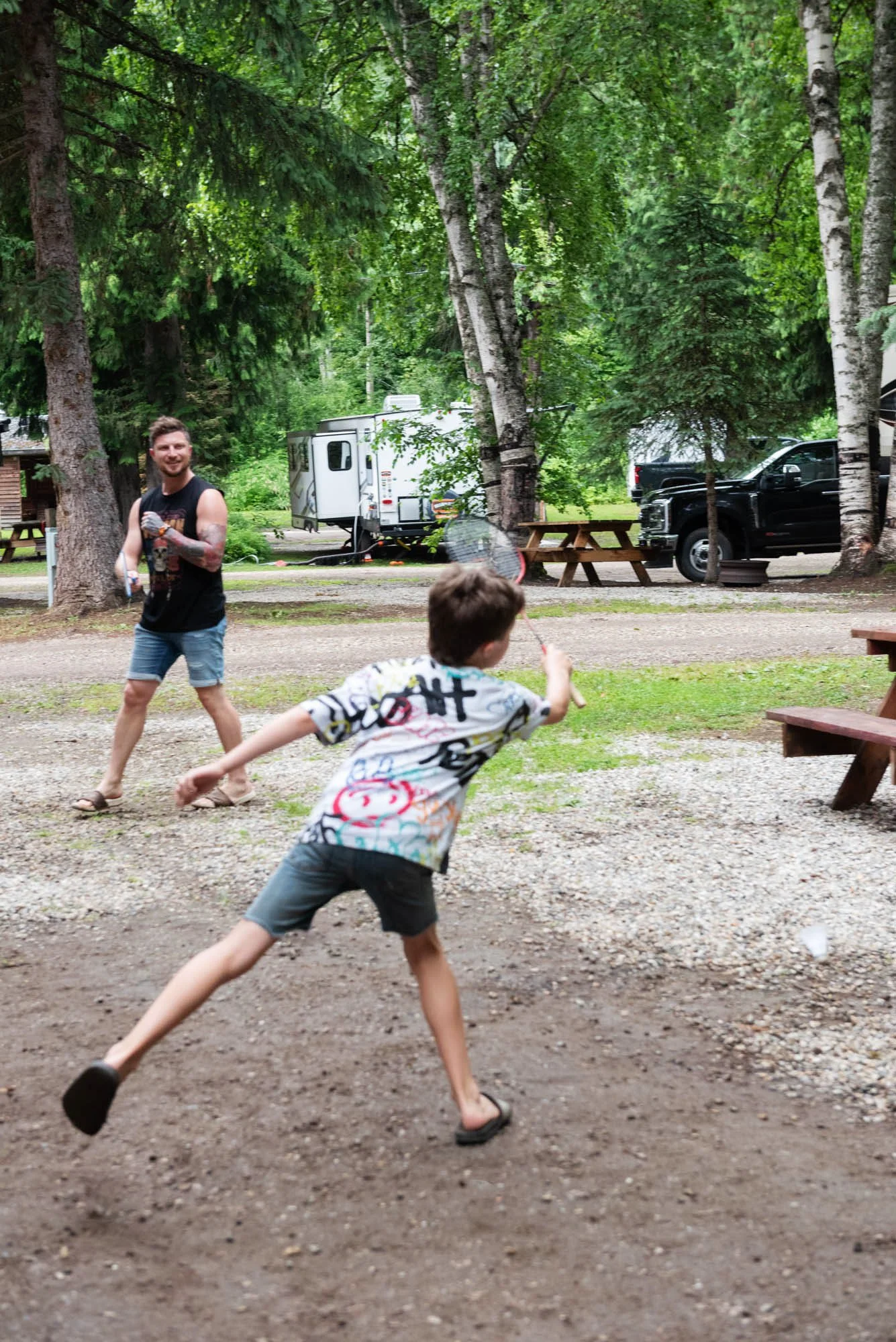 Two children playing outdoors on a dirt and gravel area in a forested park with large trees, picnic tables, a camper, and trucks in the background. One child is running with a colorful shirt, and the other is chasing with a racquet.