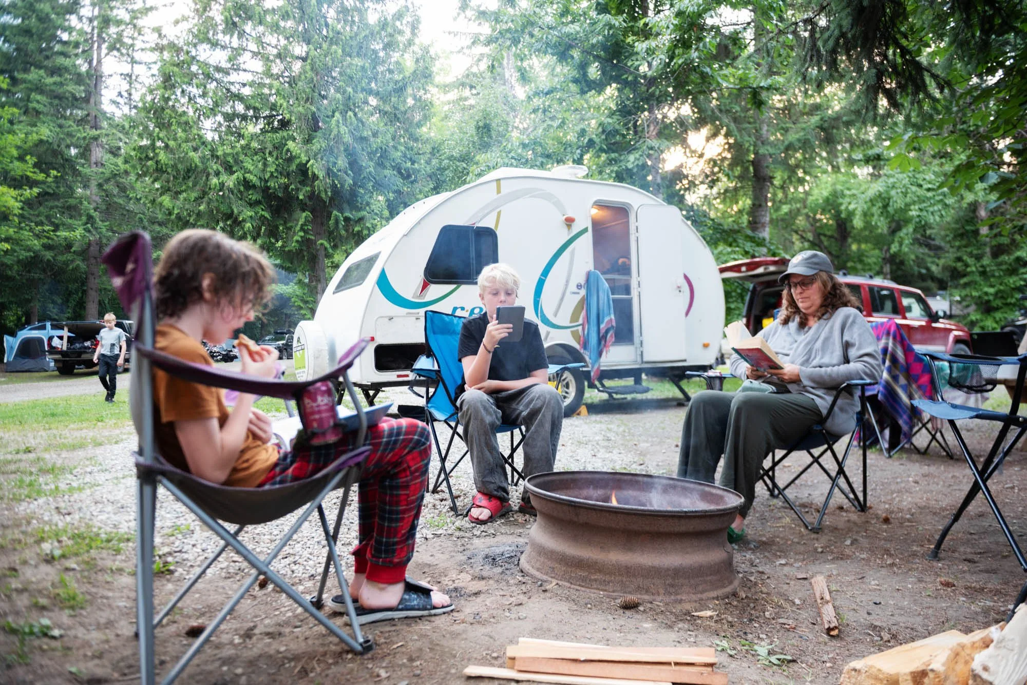 A group of four people sitting around a campfire in a wooded campsite with tents and RVs in the background. One child is eating, another is looking at a tablet, and an adult woman is reading a book.