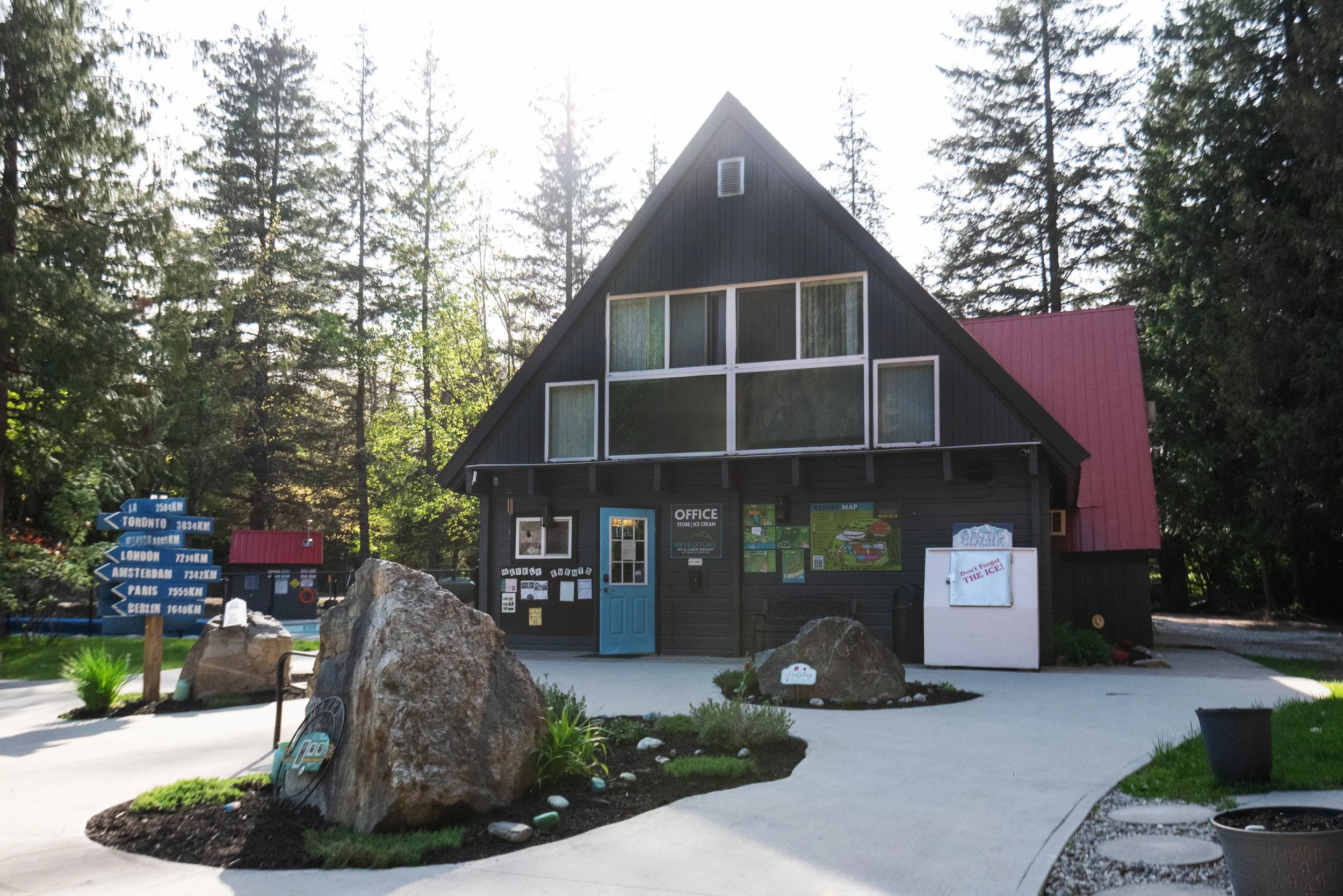 A black A-frame building with large front windows and a blue door serves as an office at a resort. There are trees surrounding the area and directional signs pointing to various international cities. Rocks and landscaped plants decorate the foregroun