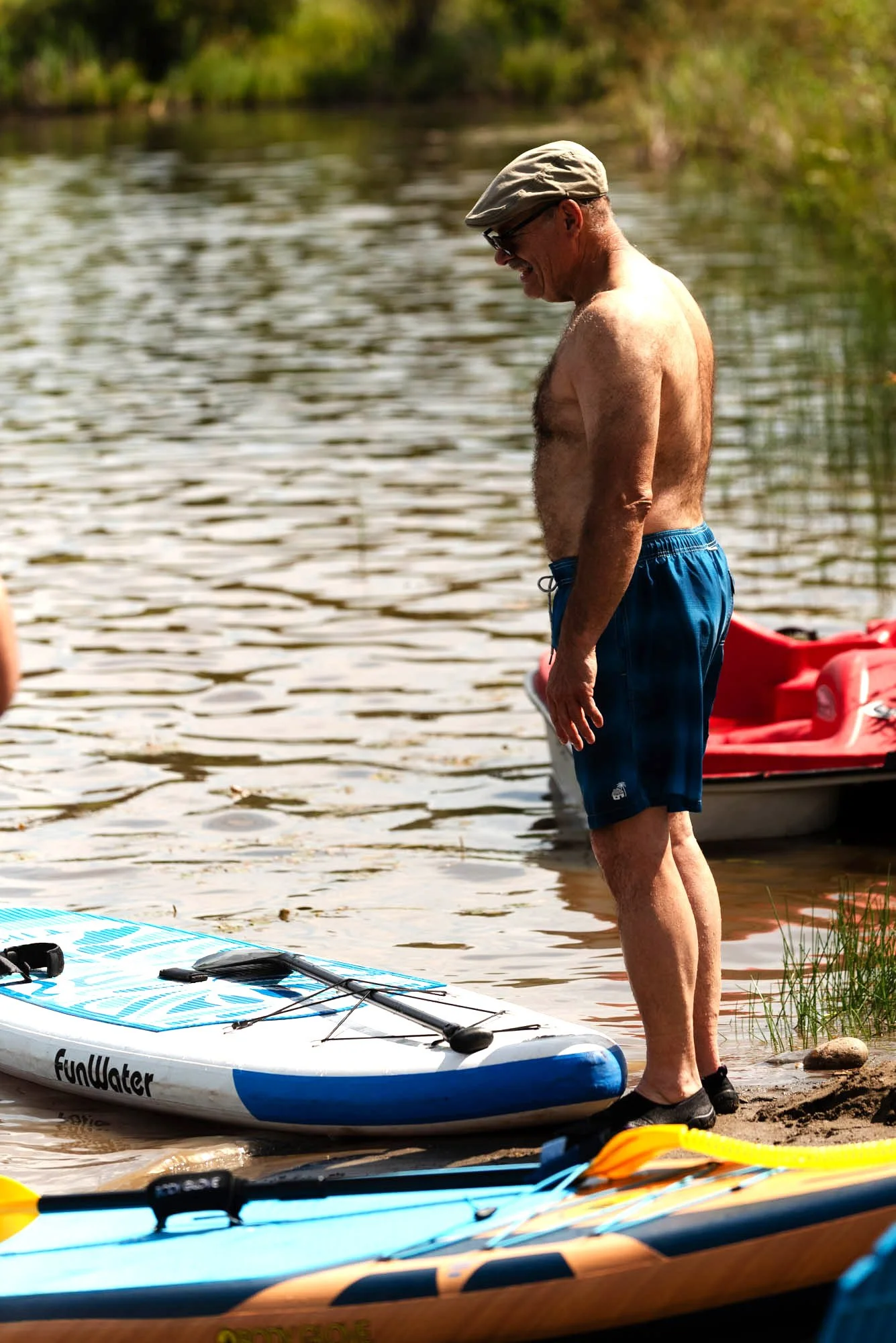 A man in blue shorts, glasses, and a beige hat standing on the shore, preparing to do water activities with paddleboards and kayaks nearby on a calm lake.