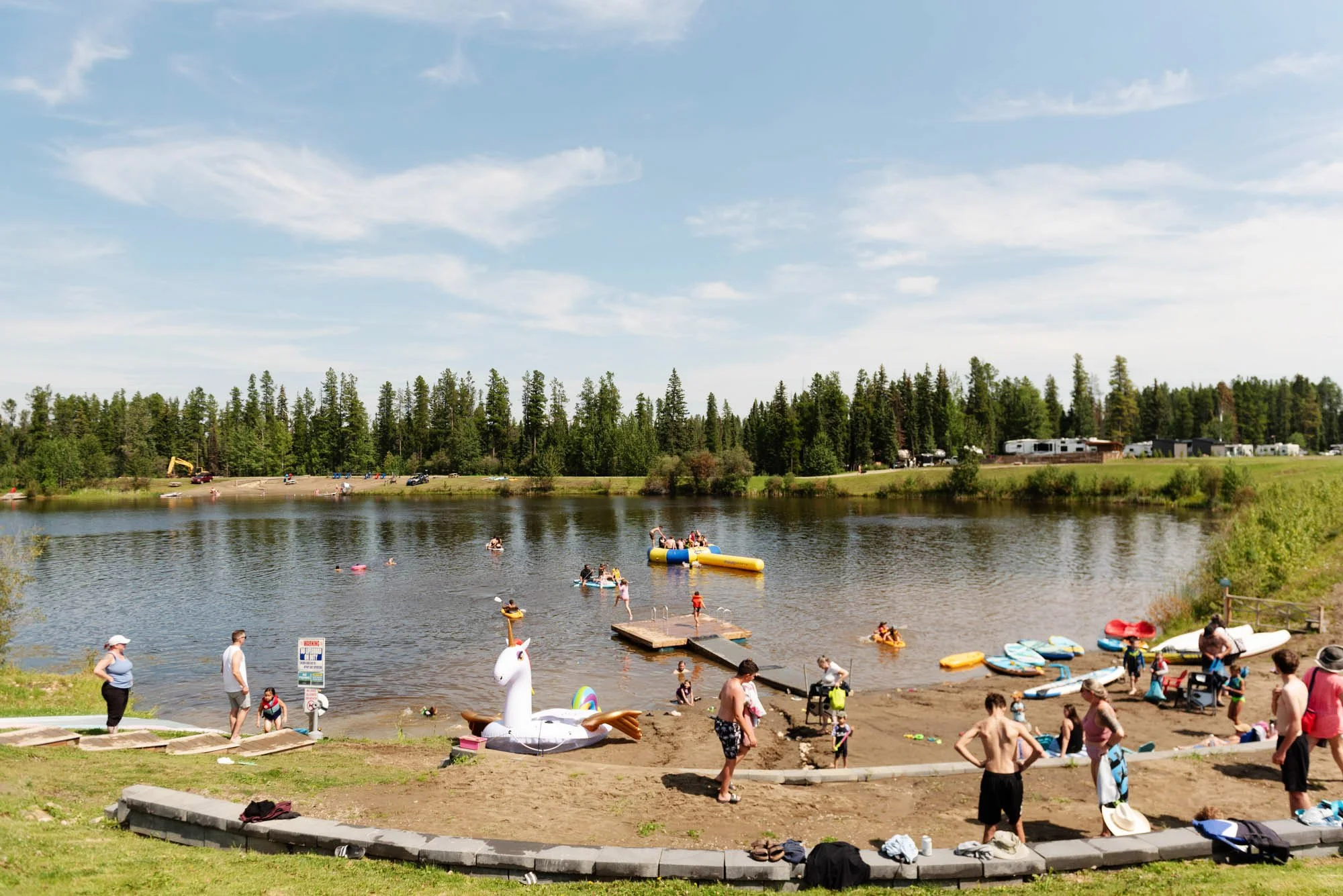 A lively lakeside scene on a sunny day with many people swimming, playing, and relaxing. Children and adults are enjoying water activities, with inflatables and kayaks visible. The background features a tree-lined shoreline, RVs, and tents, indicating a camping area.