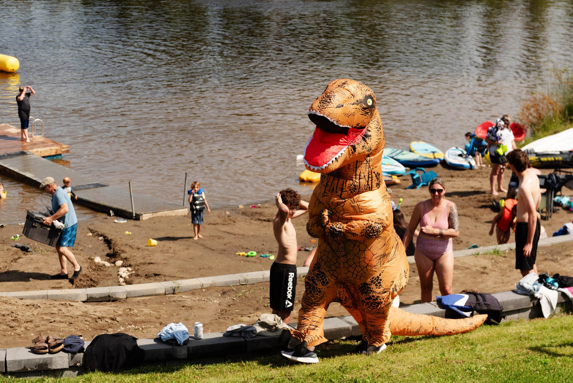 People at a beach with a person in a dinosaur costume, children playing by the water, and kayaks on the shore.