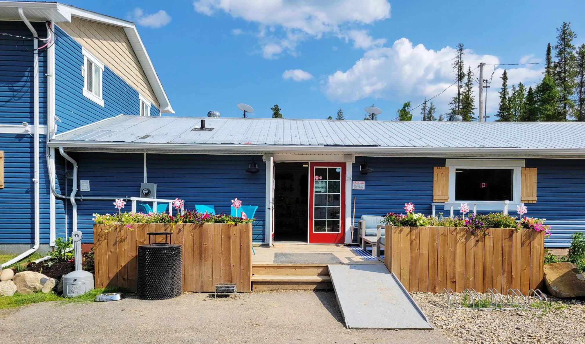 Front view of a blue house with a metal roof and red door, decorated with flower boxes, outdoor seating, and a wheelchair ramp under a blue sky with clouds. Trees and power lines are in the background.