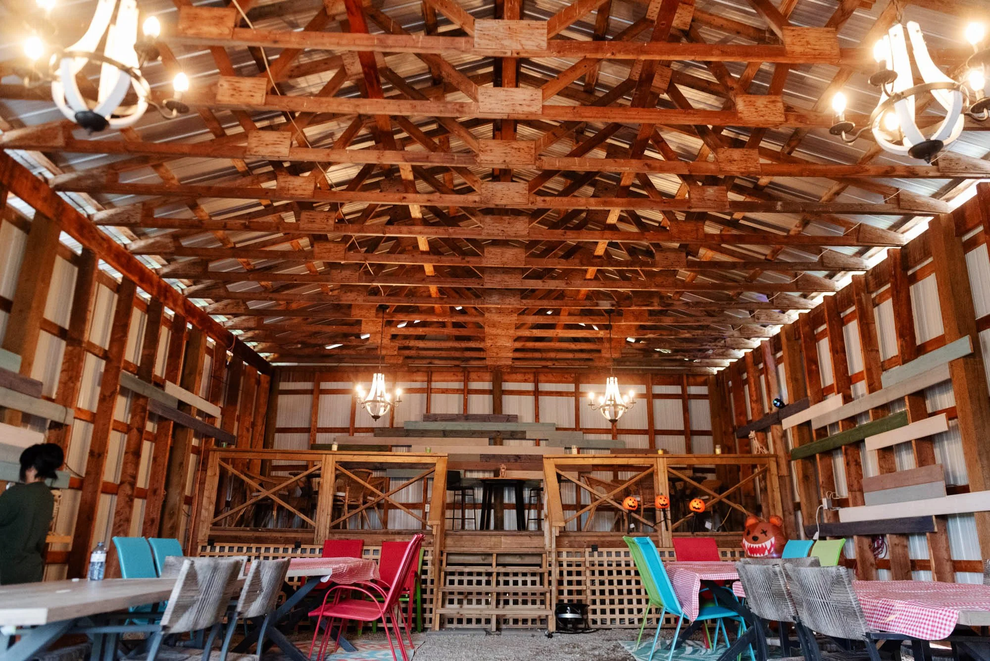 Inside a rustic barn with wooden beams and metal roofing, decorated for Halloween with pumpkin lanterns. Colorful chairs and tables are set up for an event.