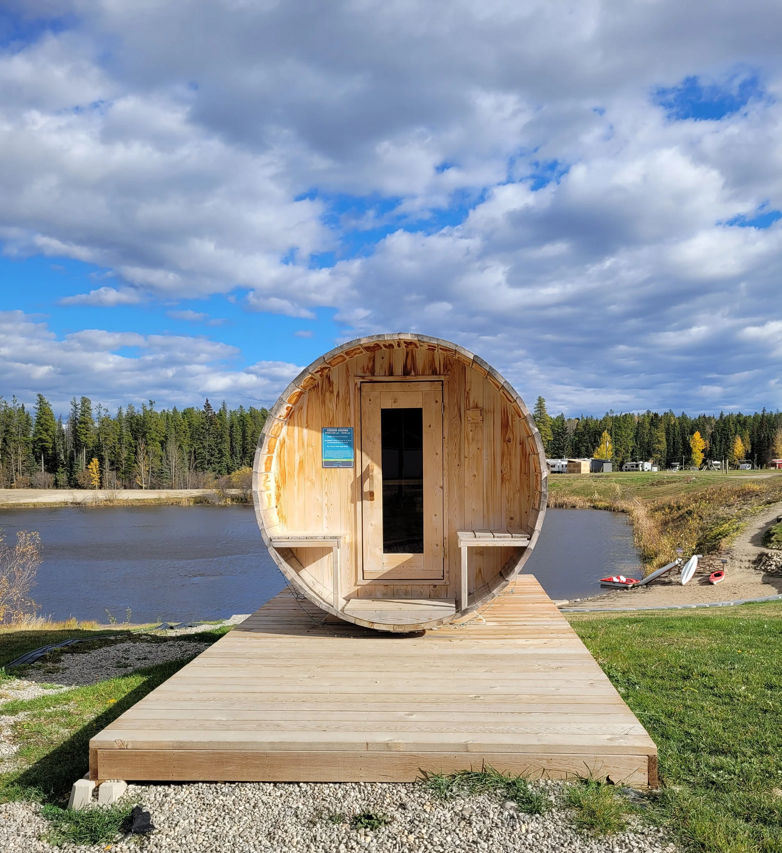 A small, rounded wooden sauna with a door, situated outdoors by a river, with a deck and surrounded by nature and trees under a cloudy sky.