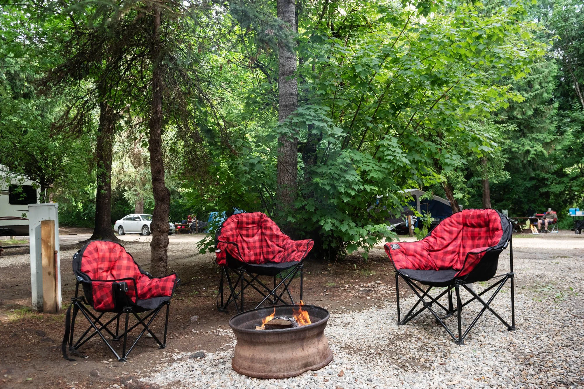 Camping chairs with red plaid fabric around a campfire in a wooded area with trees, parked cars, tents, and people in the background.