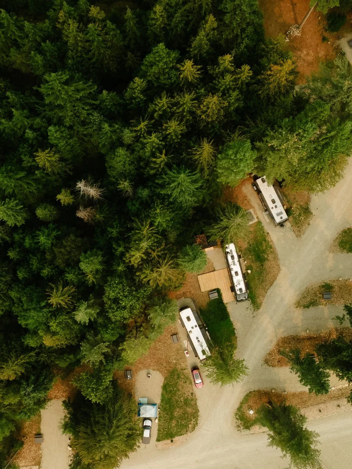 Aerial view of a wooded campground with three RVs parked near trees, and a gravel road winding through the area.