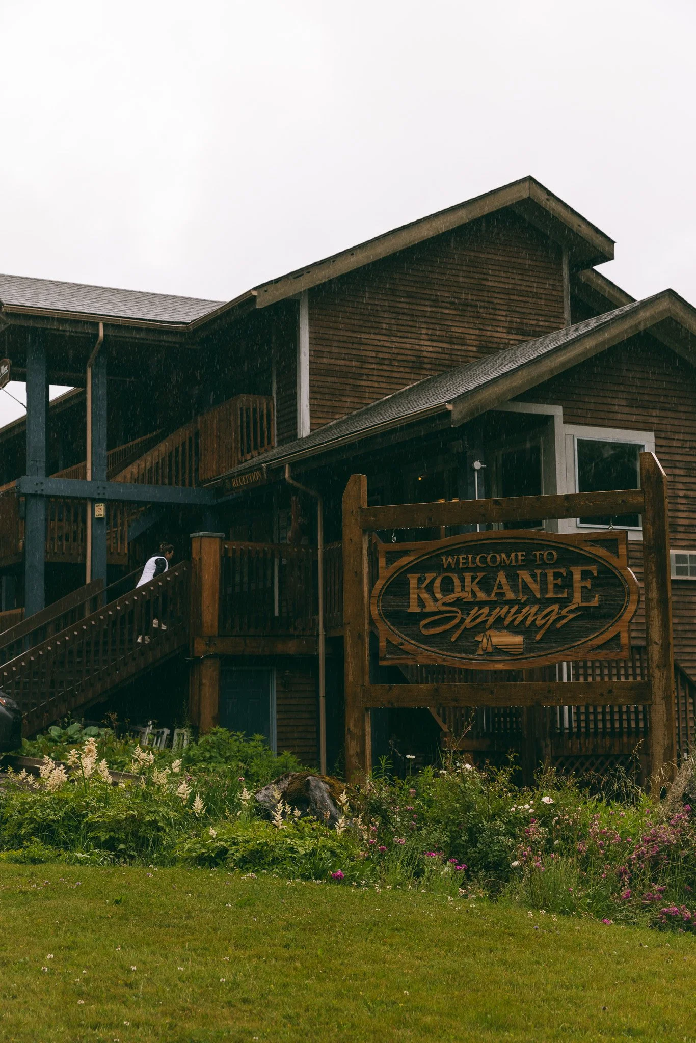 Wooden building with a sign that reads "Welcome to Kokanee Springs" surrounded by a garden with various green plants and pink flowers, and an overcast sky with rain.