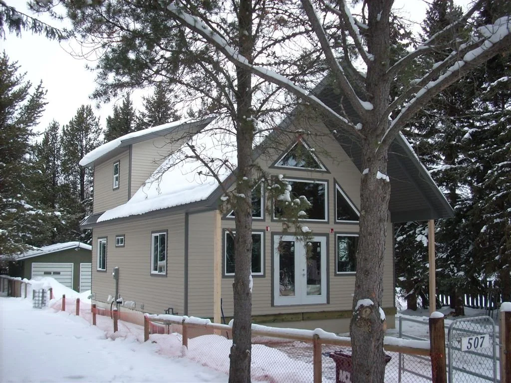 A modern two-story house in a snowy landscape, with multiple large triangular and rectangular windows, a small side building, and trees in the front yard.