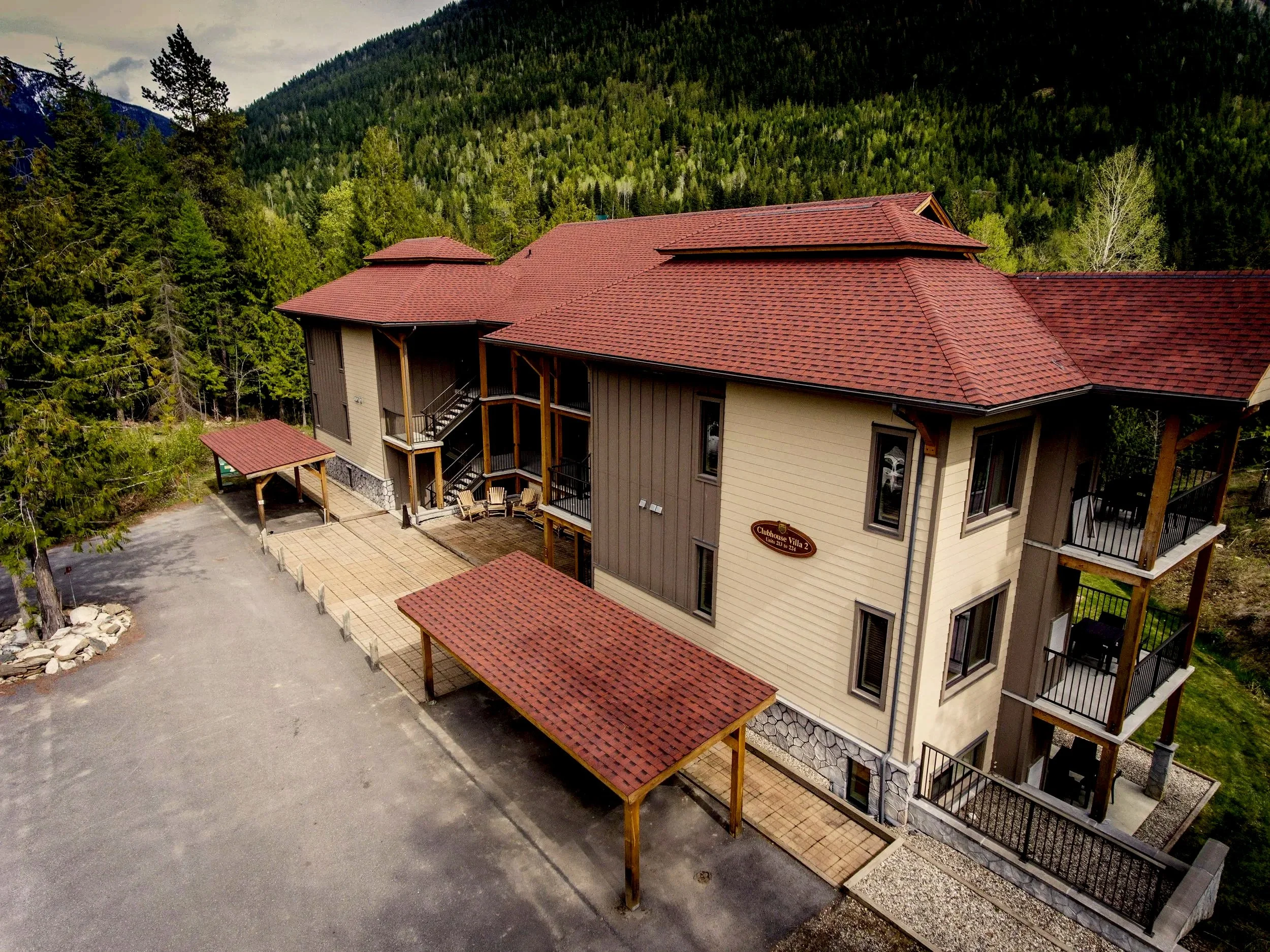 A multi-story building with a red tiled roof, nestled among dense forested mountains. The building has outdoor staircase access, balcony railings, and a sign that reads "Clubhouse Villa 2".