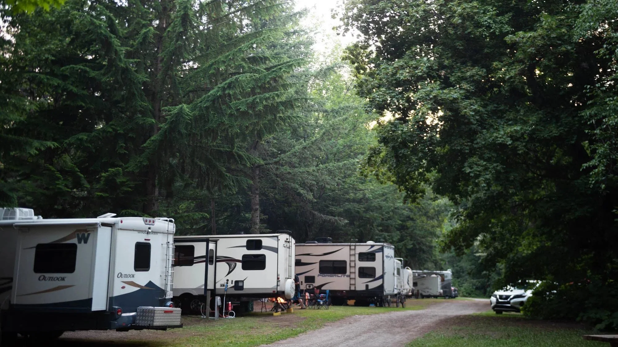 A view of a camping area with several RV trailers parked among tall green trees. The sky is partly visible through the dense foliage.