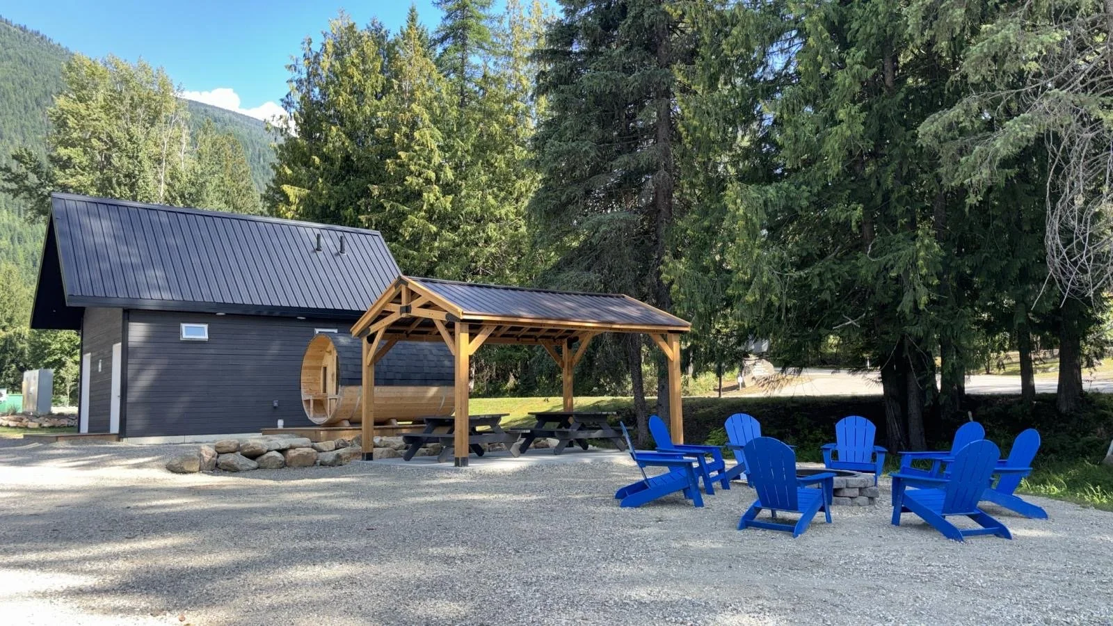 A small black building with a metal roof and wood accents, a wooden shaded picnic area with tables, and a circle of five bright blue Adirondack chairs on a gravel surface, surrounded by trees with mountains in the background.