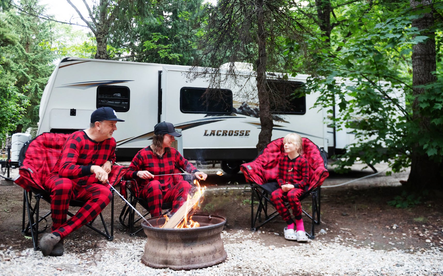Family sitting around campfire near an RV in the woods, roasting marshmallows. They are all wearing matching red plaid pajamas.