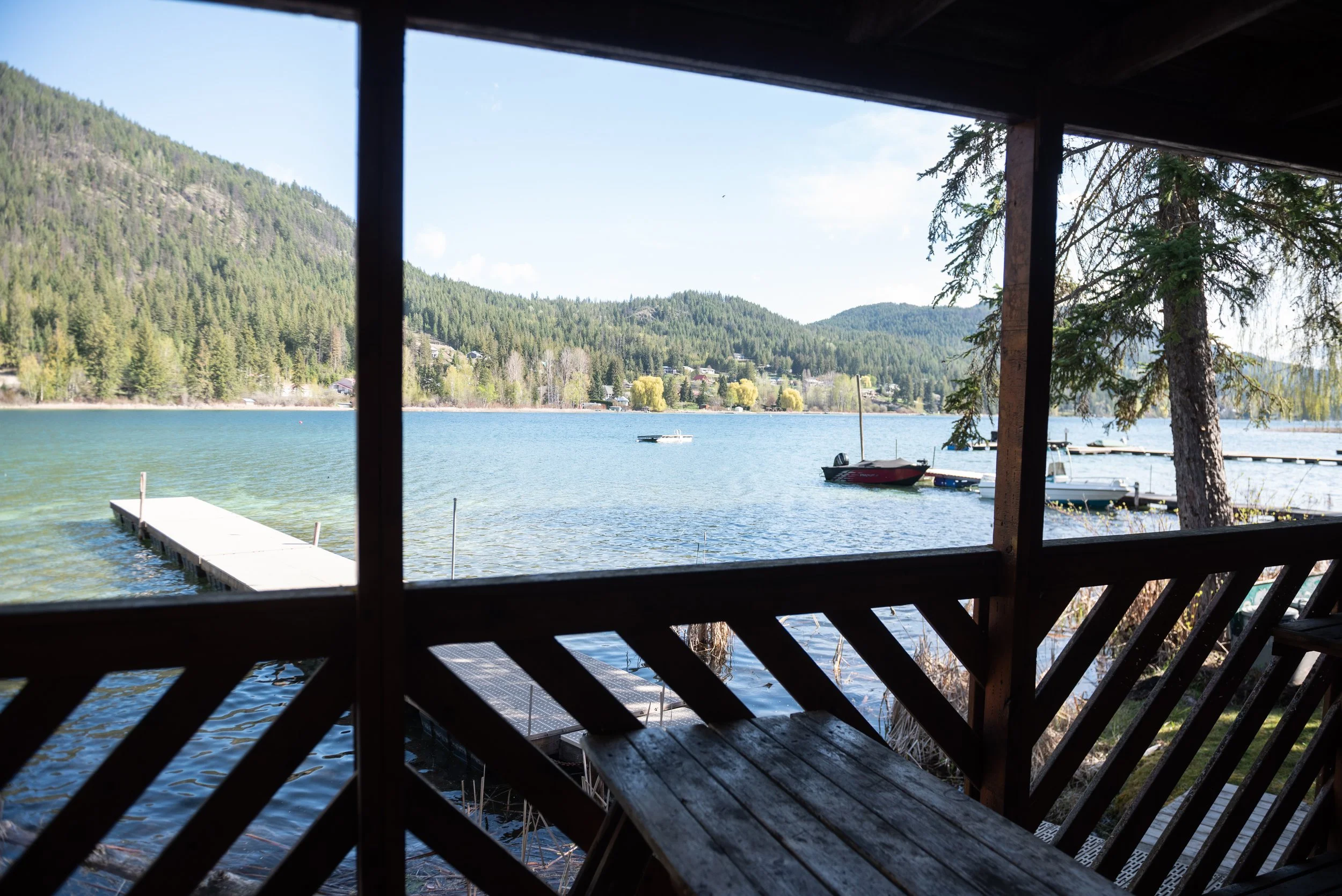 View of a lake with calm water, boats docked at a pier, surrounded by green forested hills, seen from a wooden porch or balcony with railing and posts.