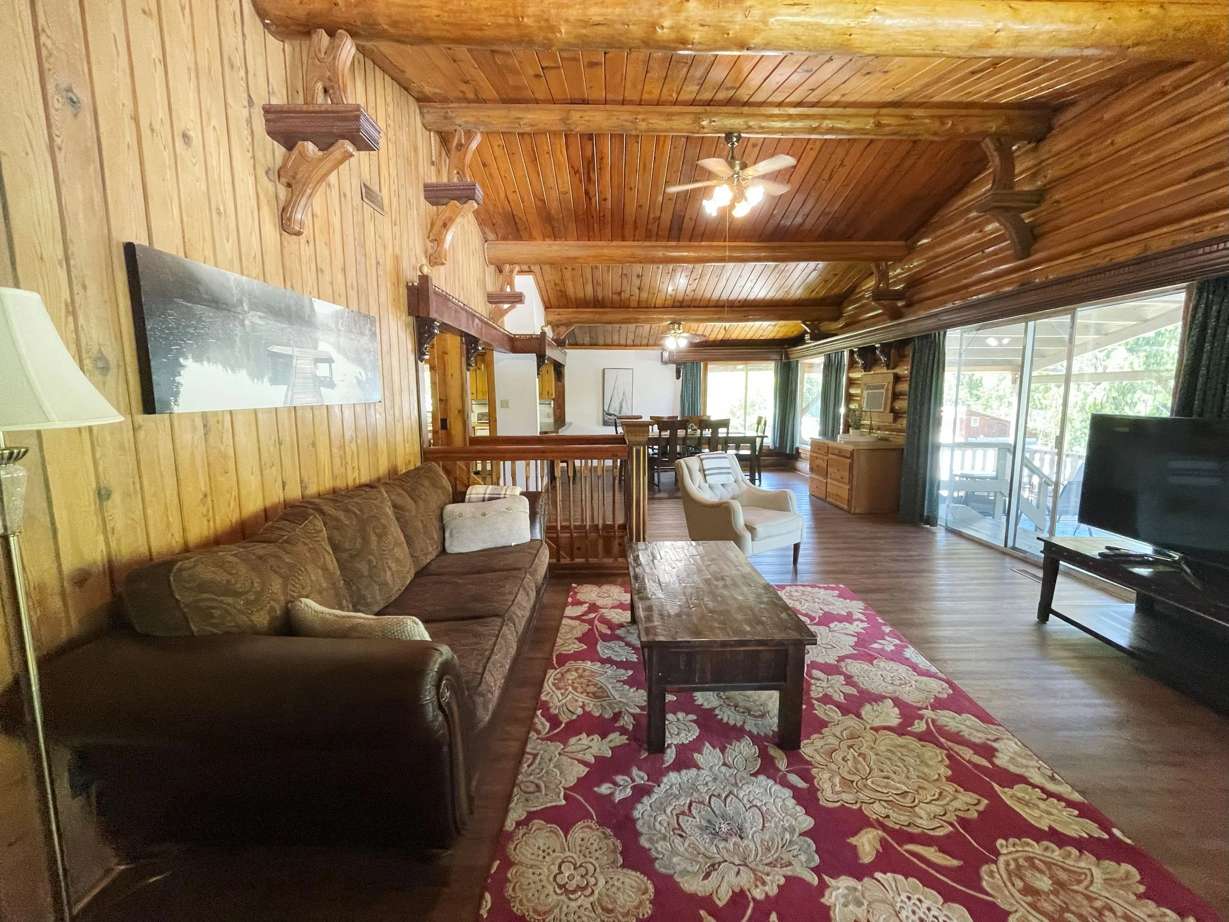 A living room with wooden walls and ceiling, featuring a brown sofa, a white armchair, a wooden coffee table, a red and gold floral rug, a large flat-screen TV, and sliding glass doors with green curtains that lead to an outside area with trees.