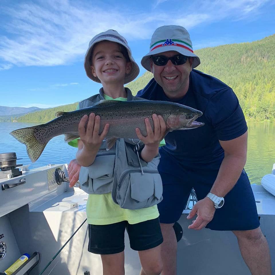 A child and an adult posing on a boat with a fishing rod, holding a large fish. They are outdoors with water and green hills in the background, under a partly cloudy sky.