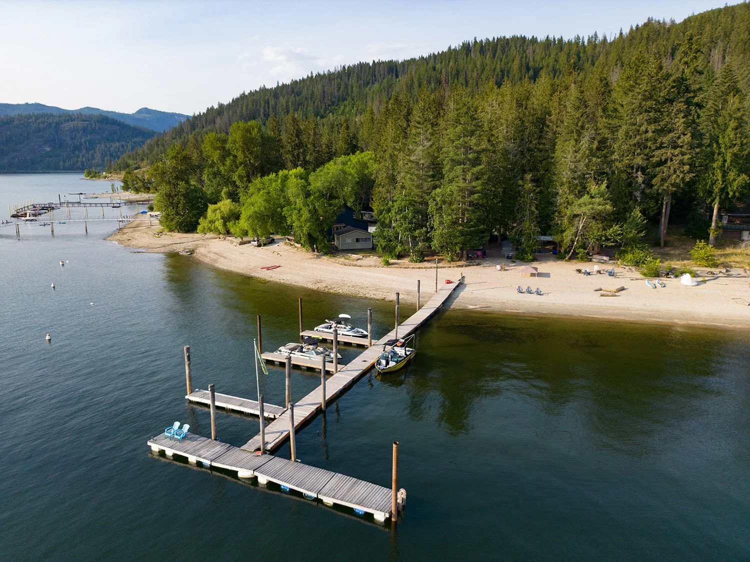 Aerial view of a lakeside dock with boats, a sandy beach, lush green trees, and forested hills in the background.
