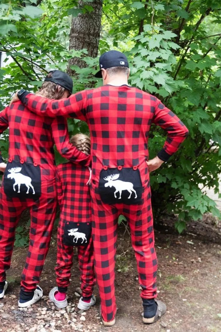 A family of three dressed in matching red and black plaid pajamas with moose designs on the back, standing outdoors near a tree with green leaves, seen from behind.