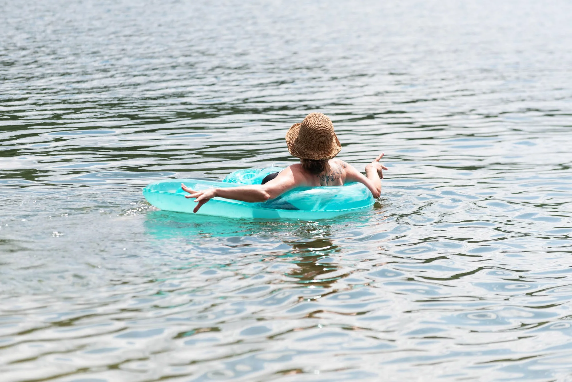 A person floating on a blue inflatable in a lake, wearing a wide-brimmed hat and black swimsuit, pointing with one hand.
