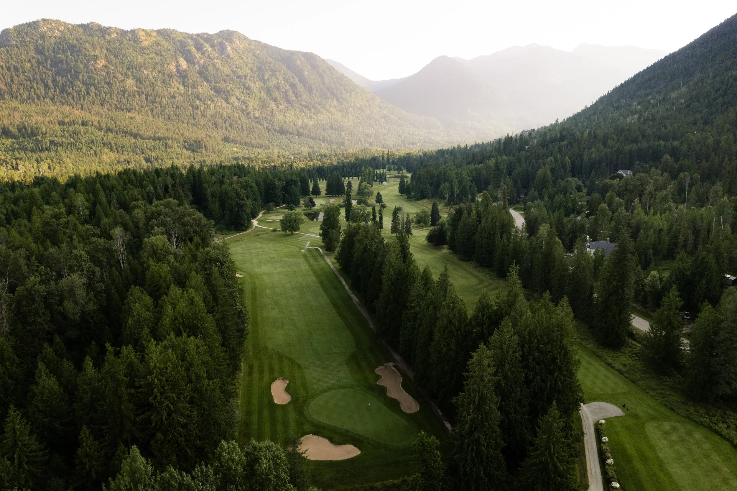 A scenic view of a golf course surrounded by dense green forest and mountains in the background. The golf course features several sand traps and a winding cart path.