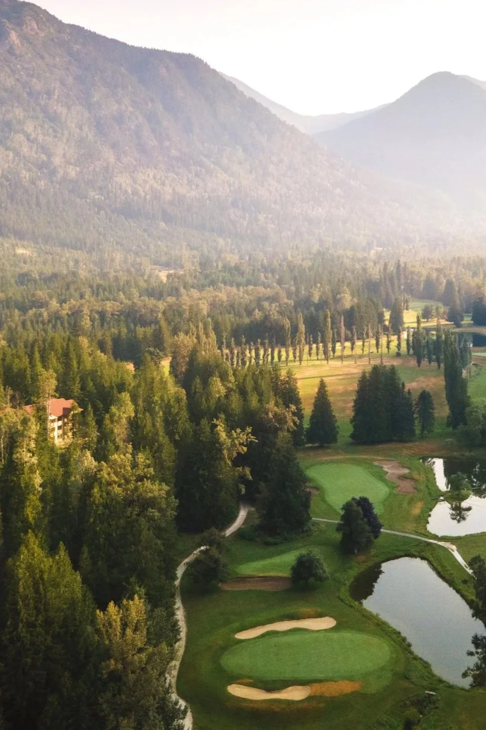 A scenic view of a golf course surrounded by tall trees, with mountains in the background and a small lake near the green.