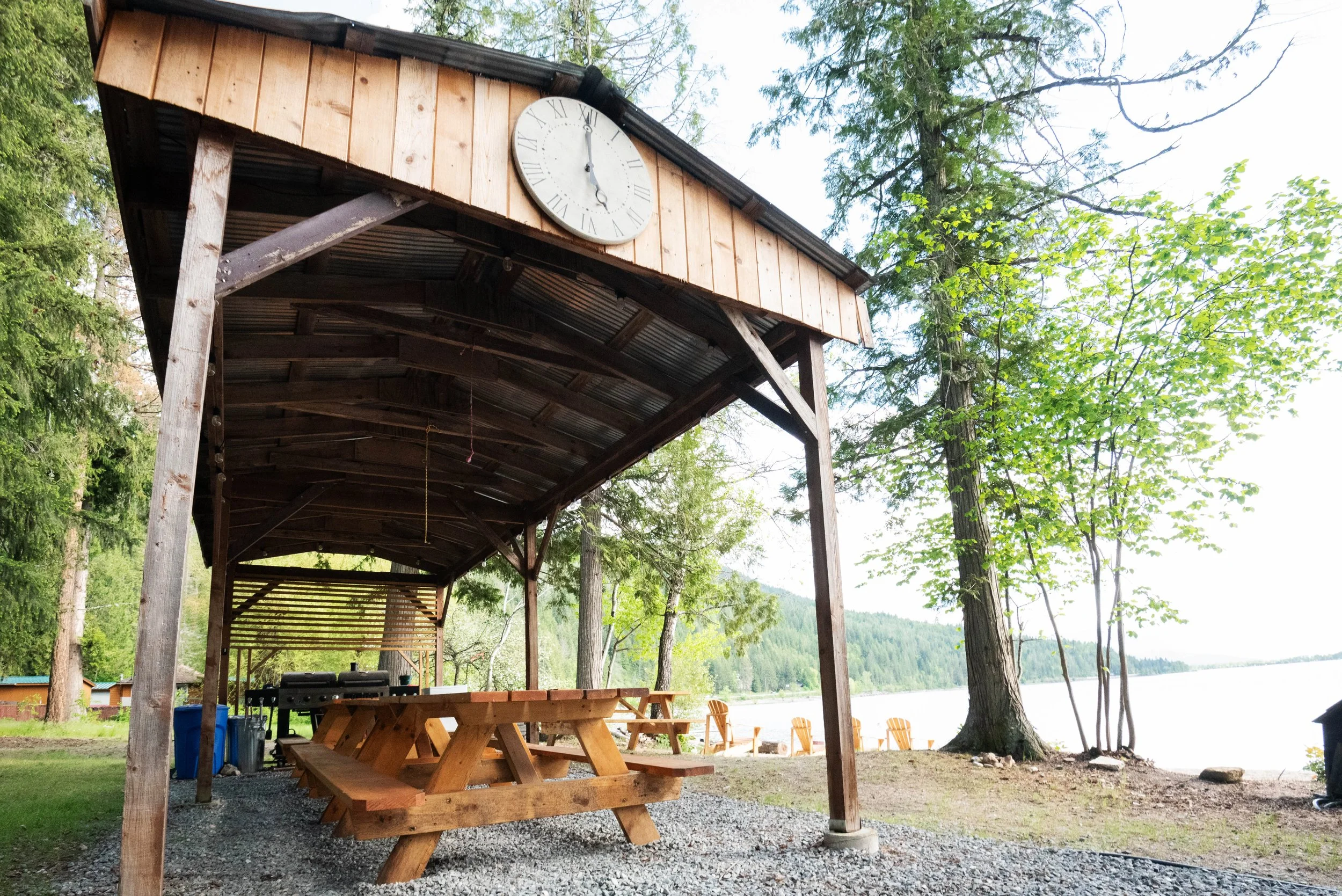 An outdoor shelter with a metal roof and wooden supports, containing several picnic tables, near a lake and surrounded by trees, with a large wall clock on the front of the shelter showing 12:28.