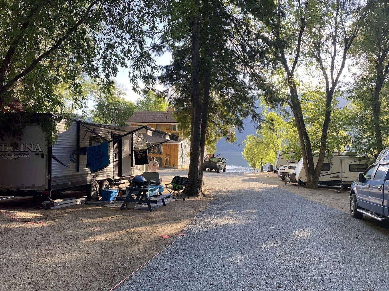 A lakeside campground with RVs and camping equipment, trees providing shade, and a view of the lake in the background.