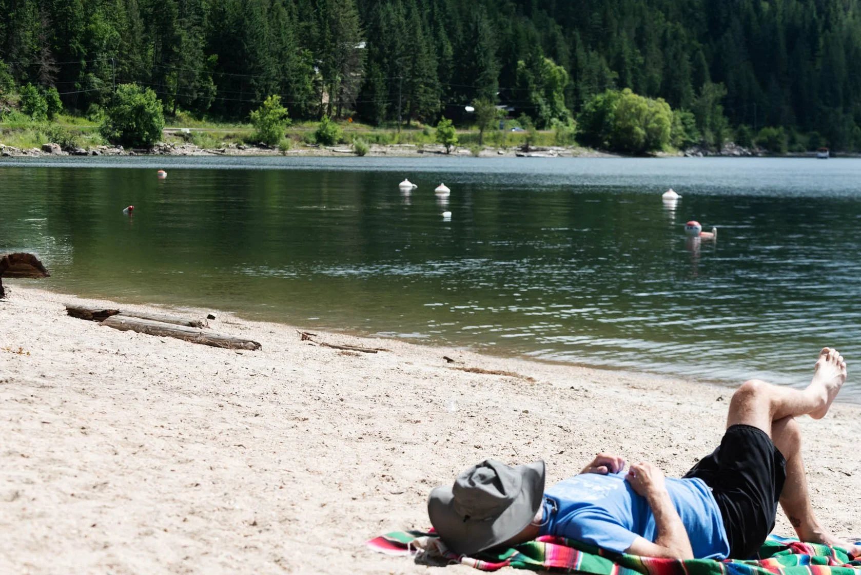 Person lying on a colorful towel on a sandy beach, with legs crossed and wearing a hat, near calm water with buoys, surrounded by green trees and forested hills.