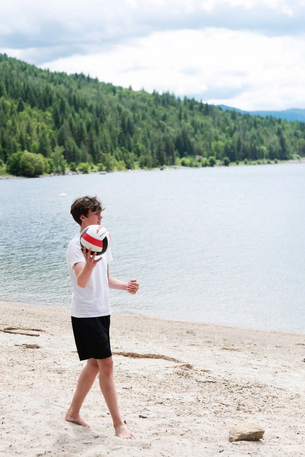 A young boy walking barefoot on a sandy beach, holding a volleyball, with a lake and forested hills in the background.