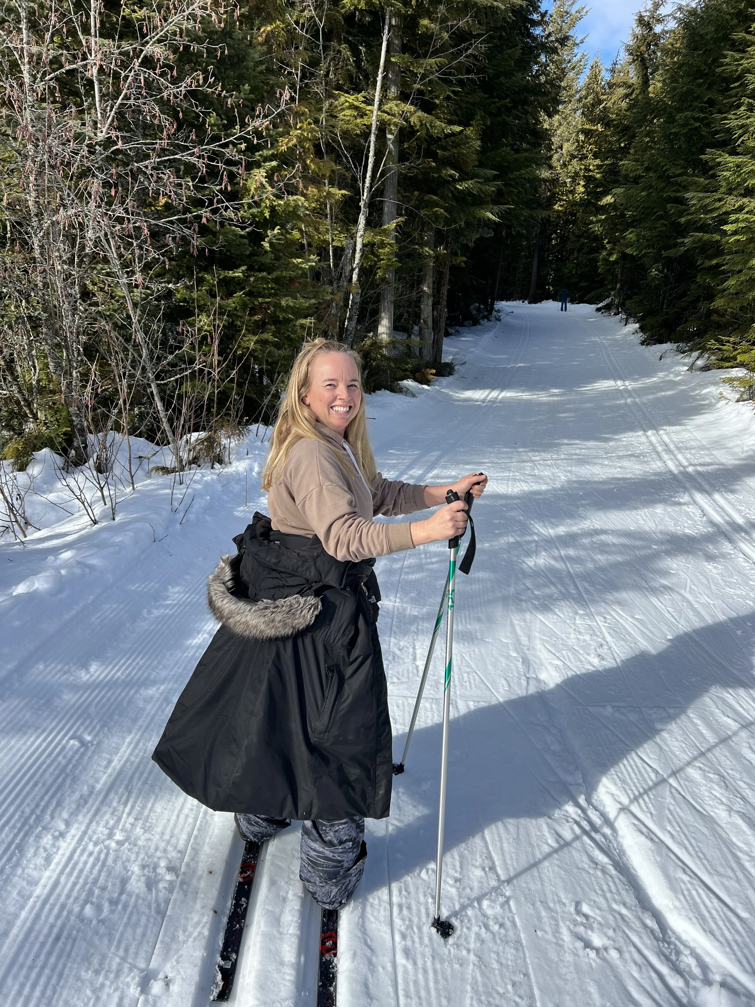 A woman smiling and holding ski poles while standing on cross-country skis in a snowy forest trail during daytime.
