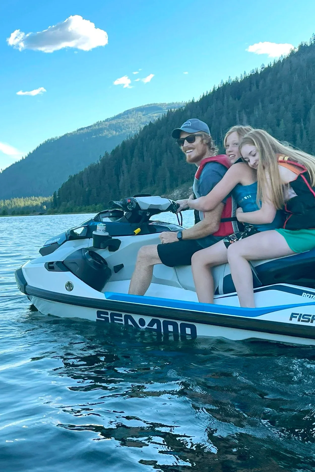Three people riding a jet ski on a lake, with mountains and a blue sky in the background.