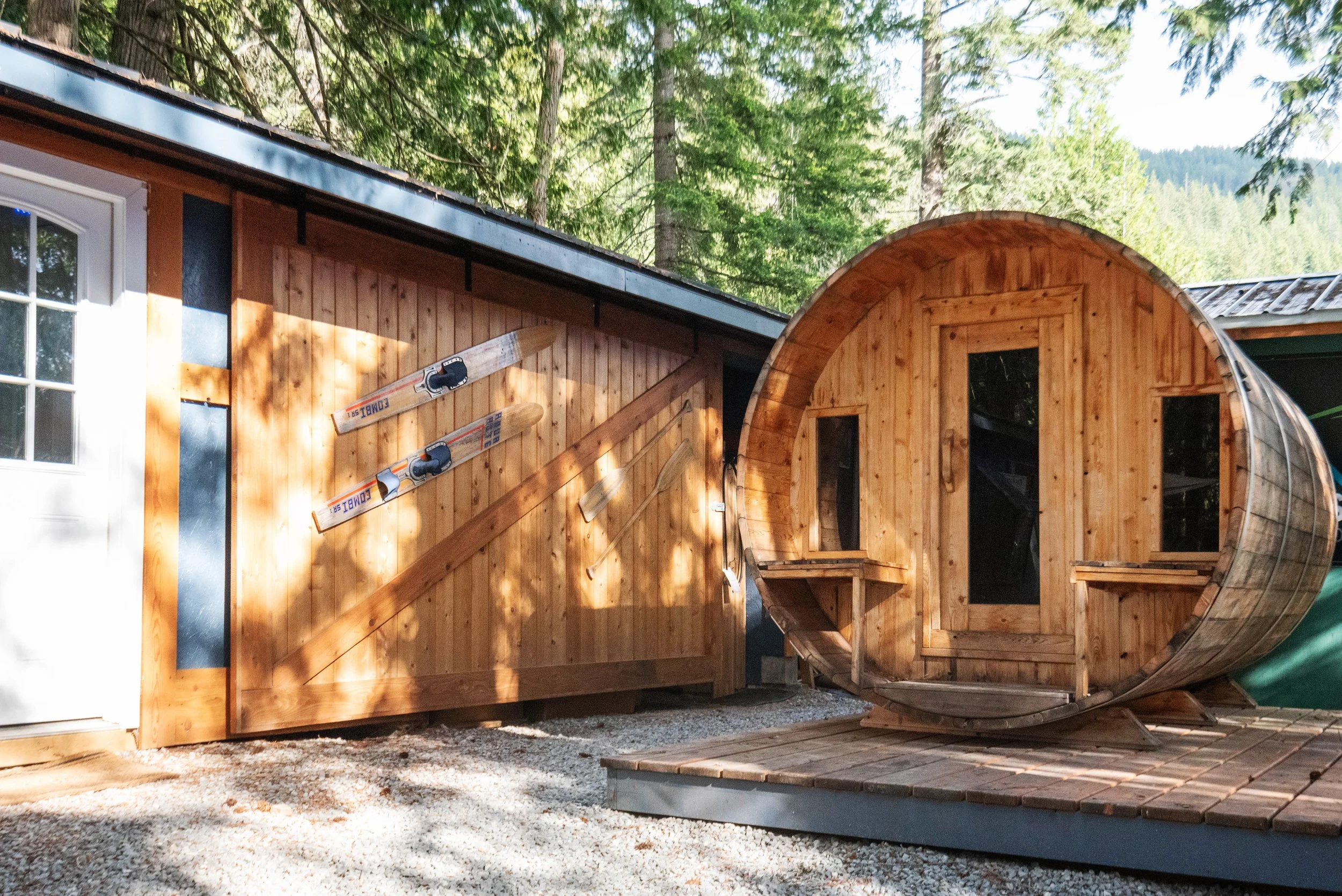 A wooden tiny house under construction in a forested area with tall trees in the background. The house has a curved roof and exterior wooden panels, and there are skis hanging on the side of the house.