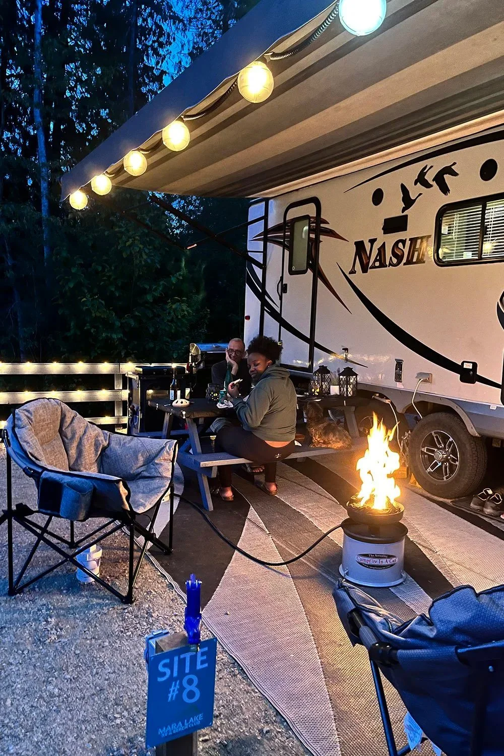 People sitting at picnic table outside of travel trailer with illuminated lanterns and a small bonfire in a metal container. Nighttime setting with trees in the background.