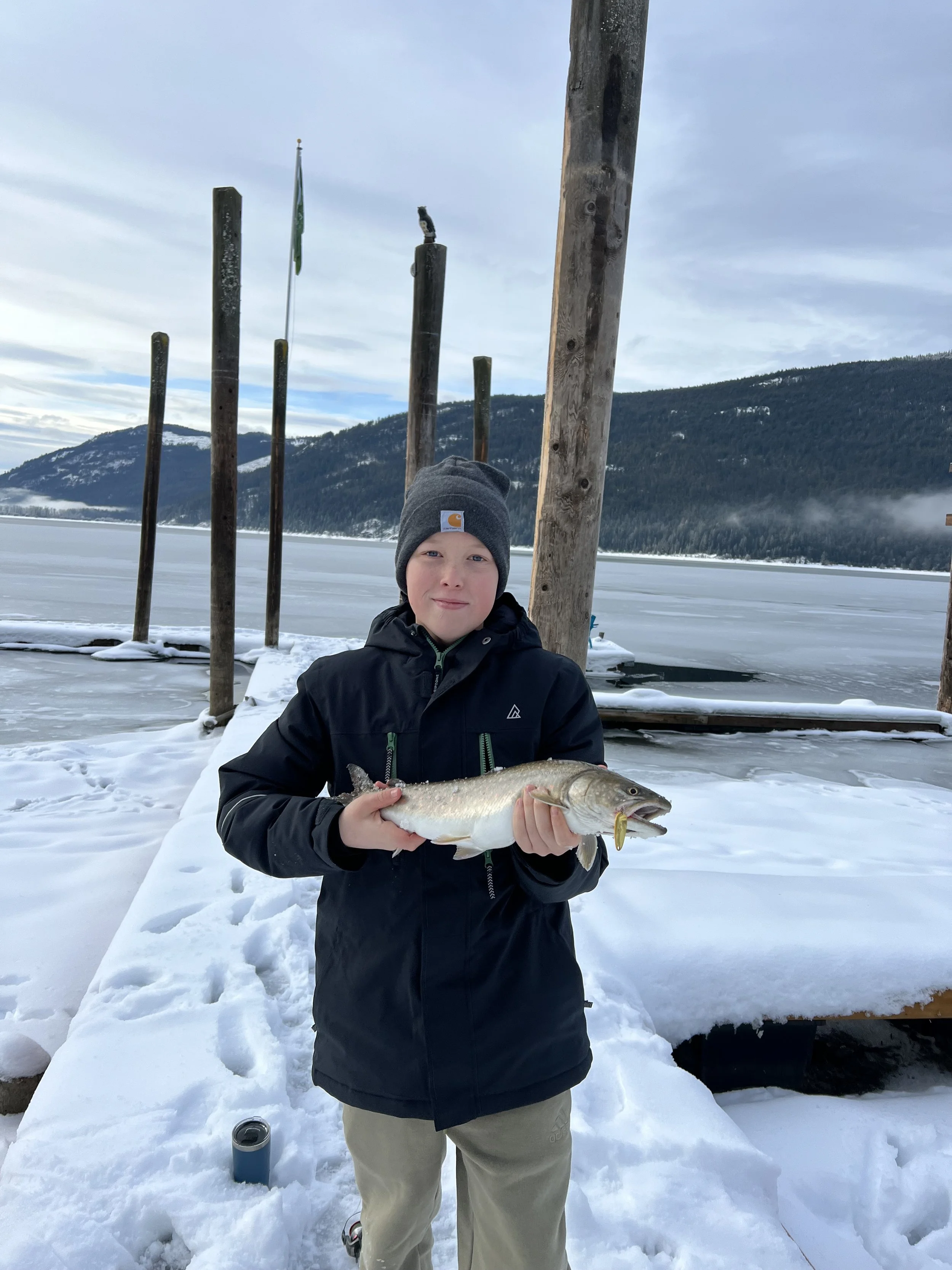 A young boy wearing a gray beanie and black jacket standing on a snow-covered dock, holding a large fish with a yellow fishing lure in its mouth. In the background, there is a frozen lake, snow-capped mountains, and a cloudy sky.
