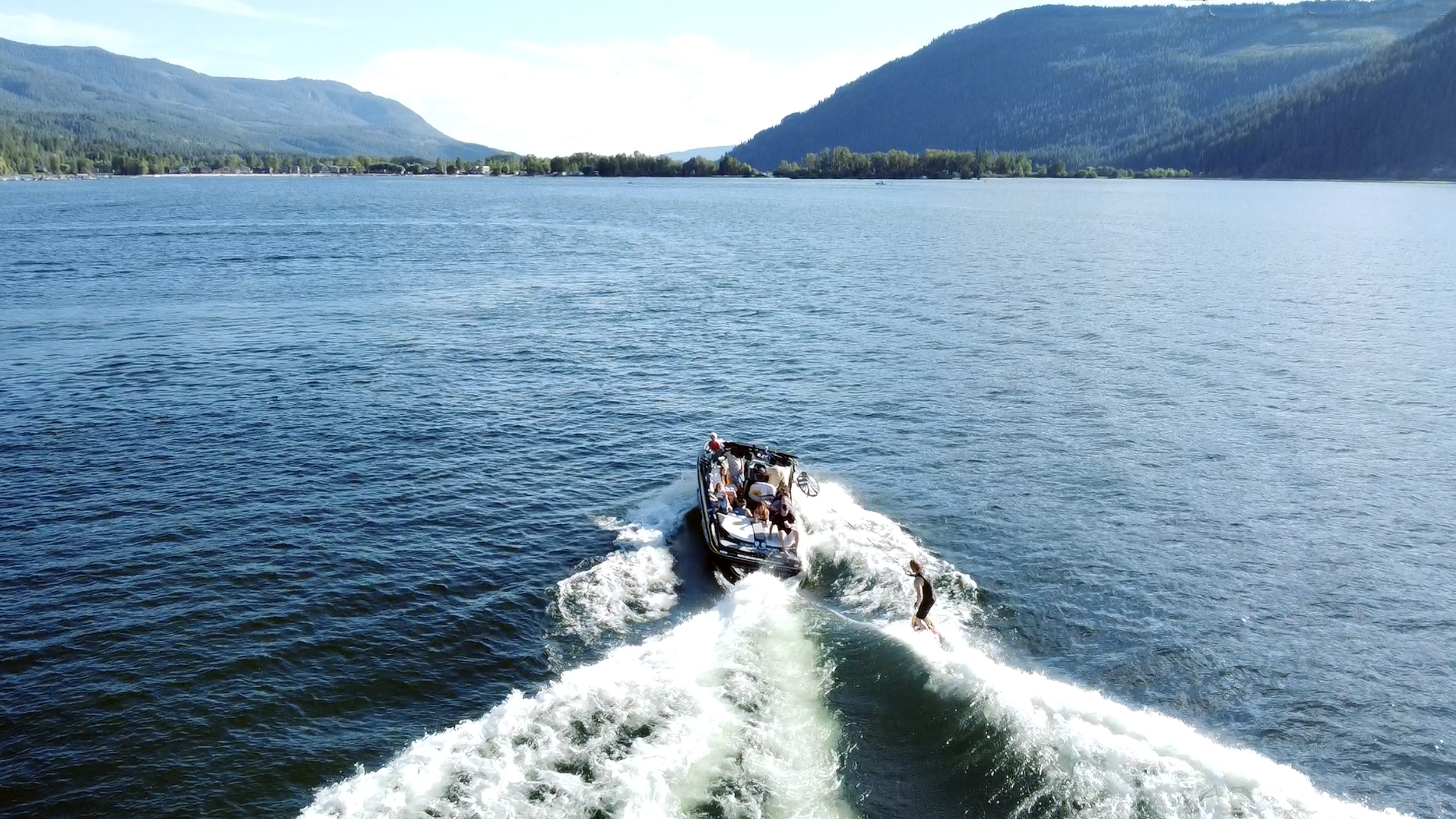 A boat is speeding across a large body of water with a person wakeboarding behind it. The water is surrounded by forested mountains under a clear sky.