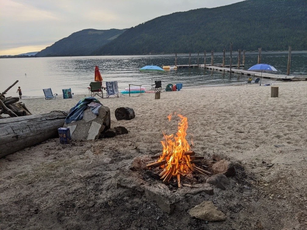 Camping scene on a beach with a campfire, beach chairs, umbrellas, a dock extending into the water, and green hills in the background
