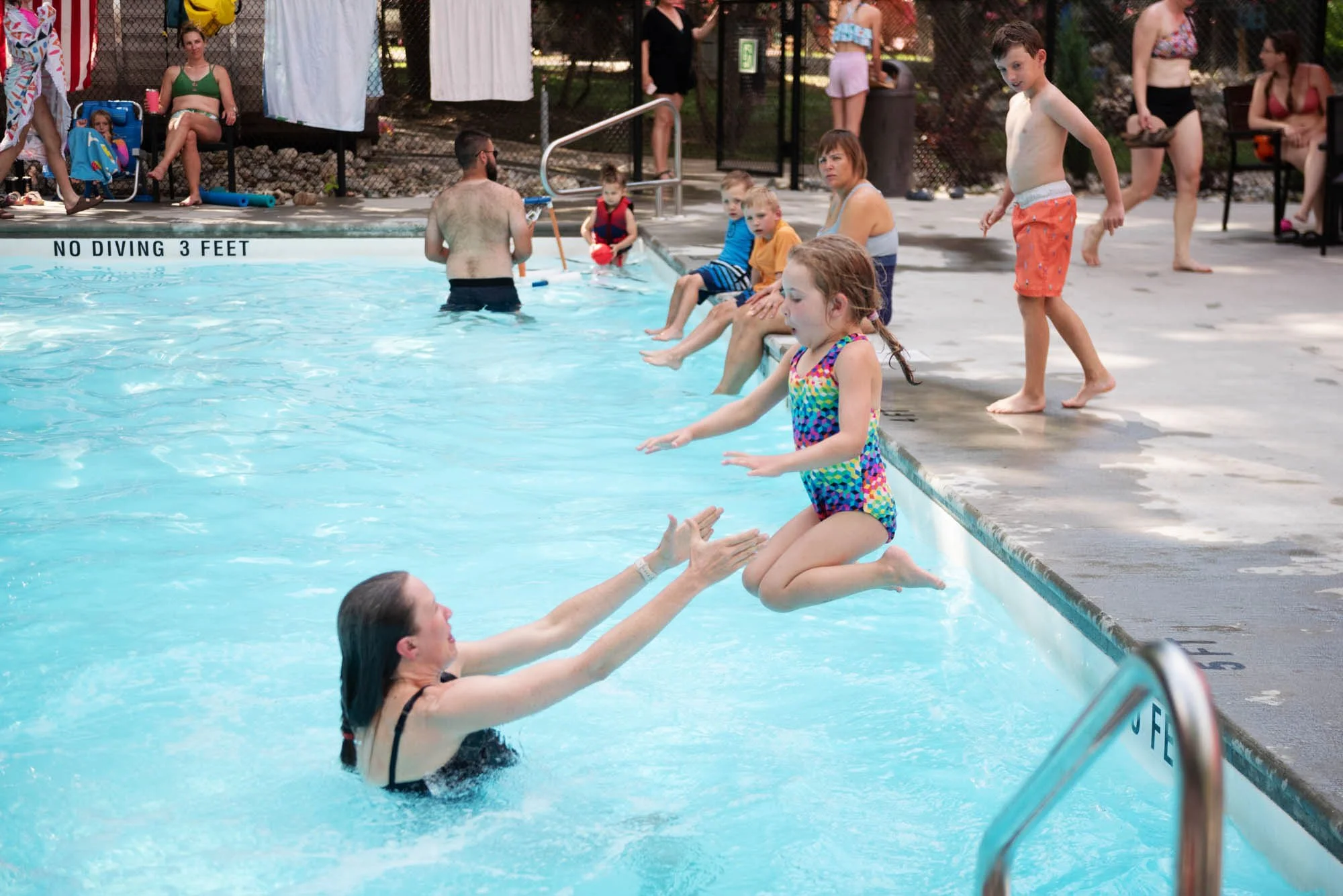 A girl in a colorful swimsuit is being handed out of a swimming pool by an adult woman with dark hair. Several children and adults are at the poolside, with some sitting on the edge and others standing nearby. In the background, there are benches, a fence, and a sign that reads 'No Diving 3 Feet'.